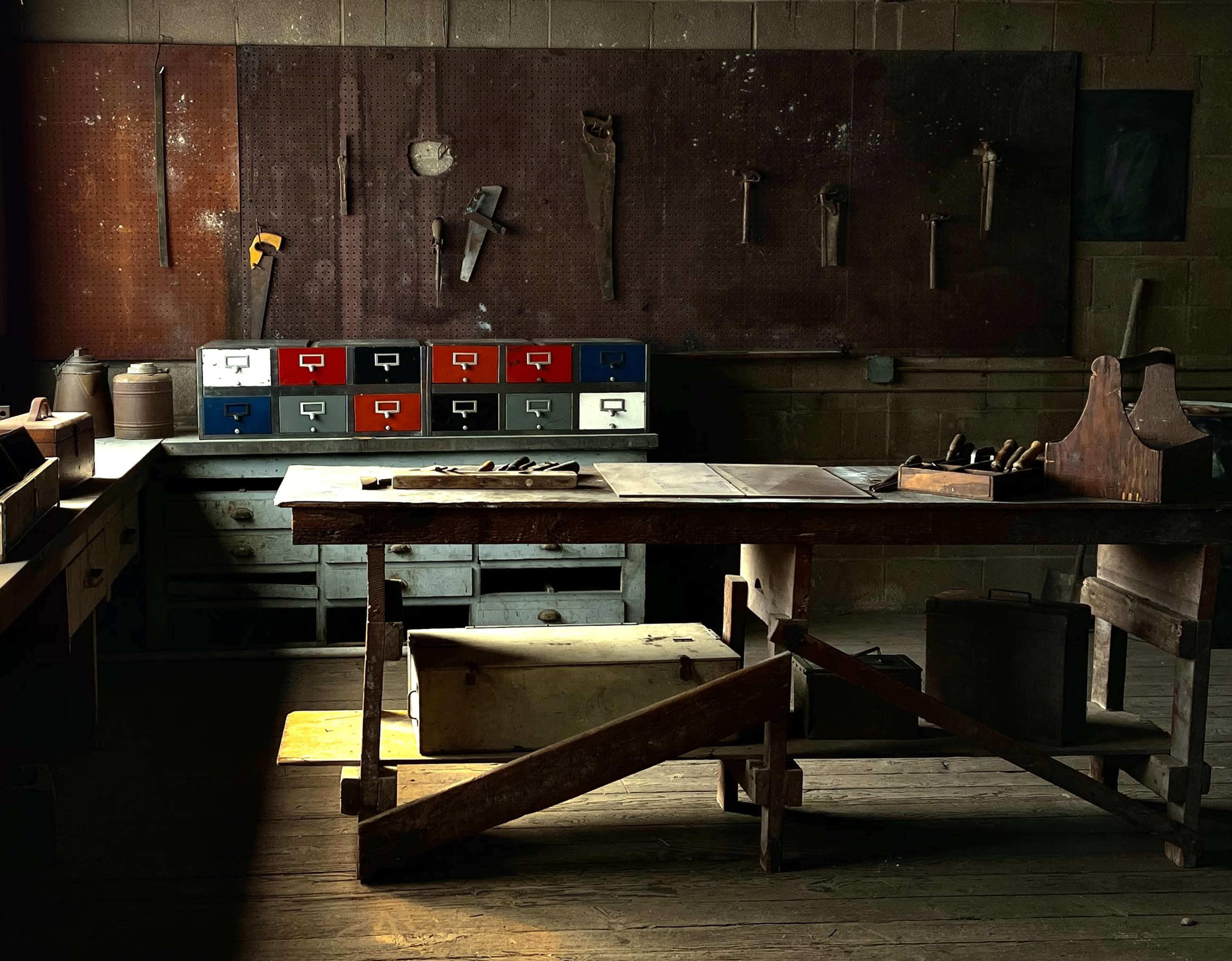 A dimly lit workshop features a large workbench, various tools hanging on the wall, and cabinets with colorful drawers.