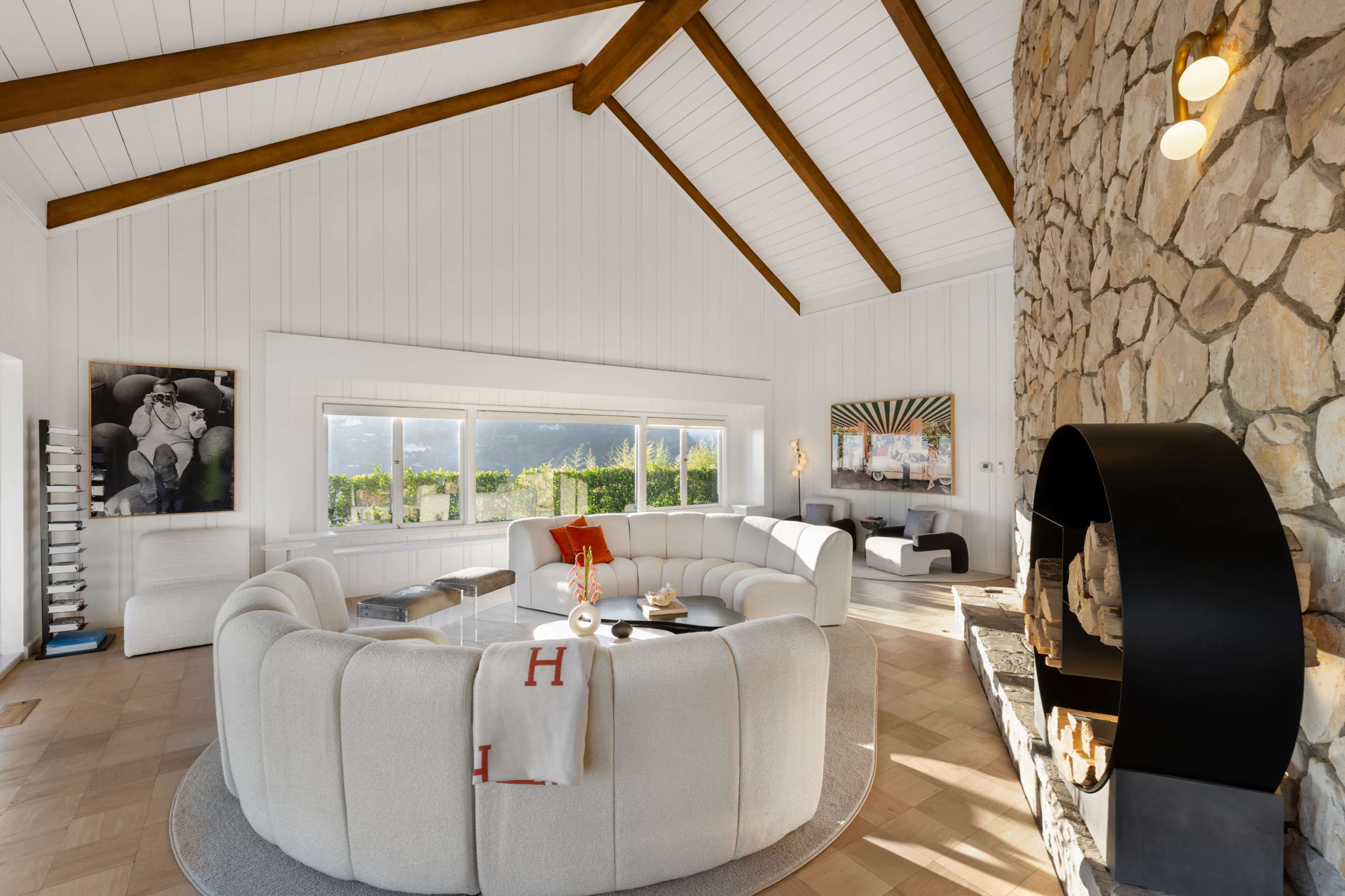 A modern living room with a high ceiling, a circular white sofa, a stone accent wall, and large windows overlooking a landscape.