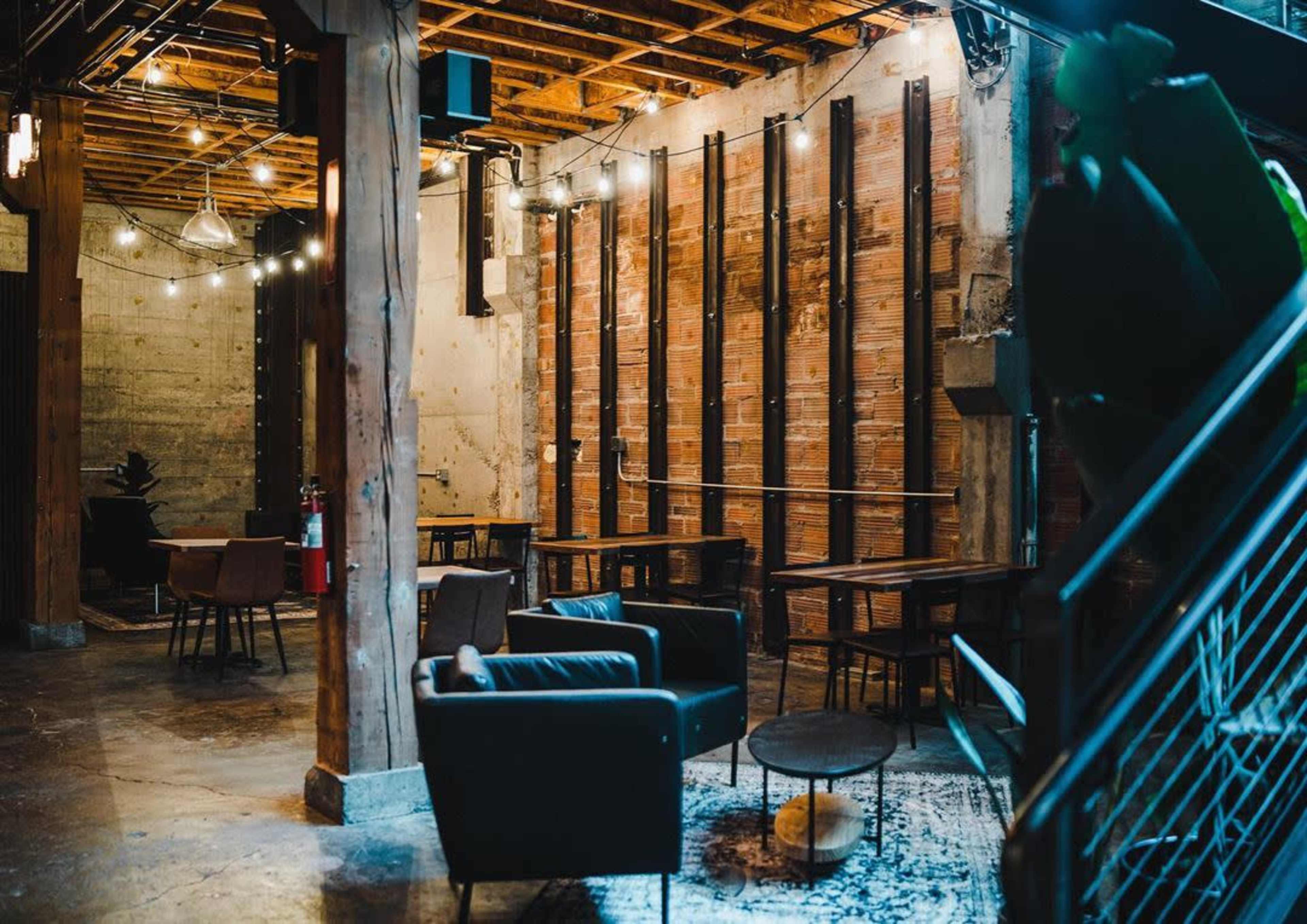 A modern café interior features wooden beams, exposed brick walls, and multiple seating areas with tables and chairs under string lights.