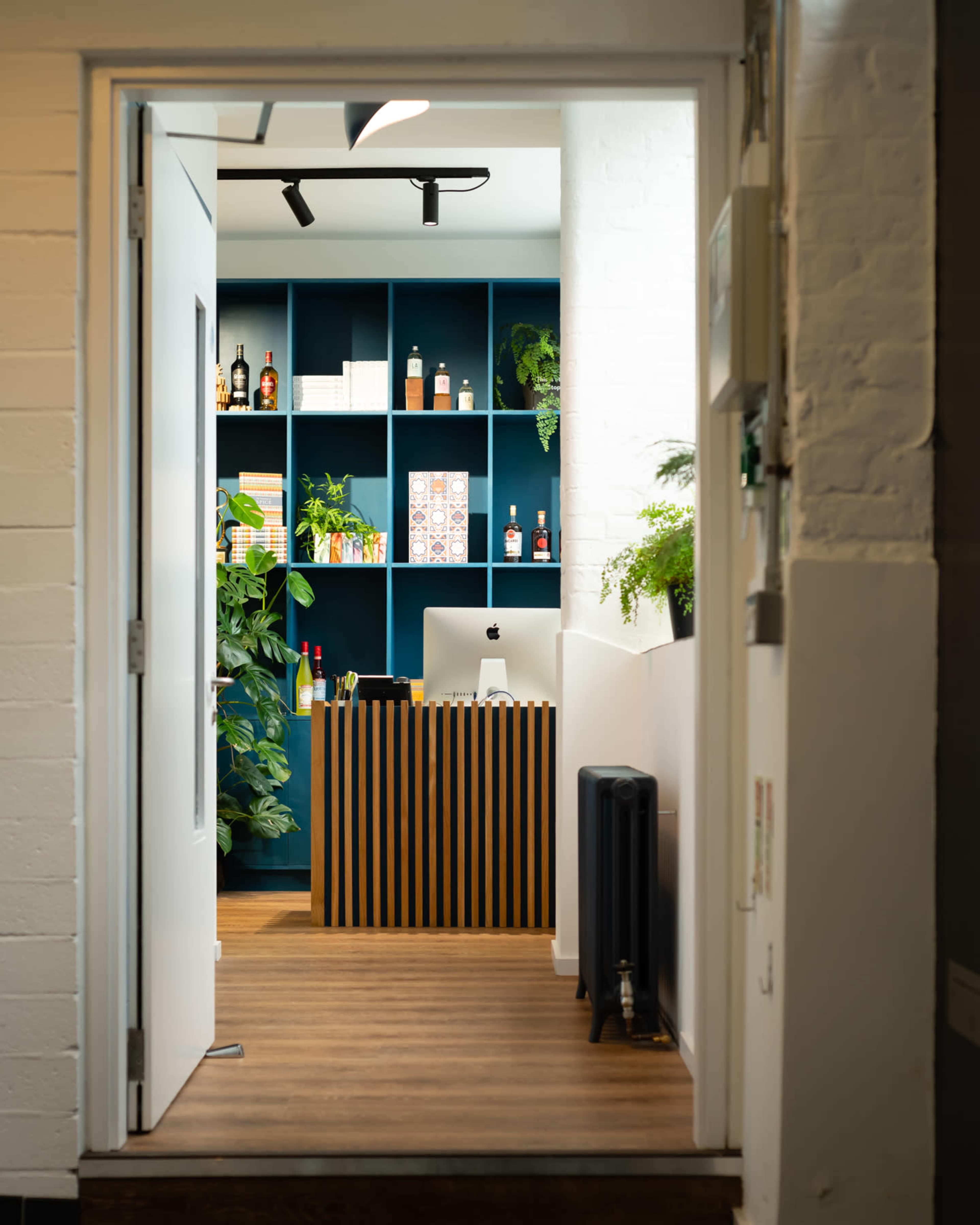 A well-designed office space features a reception area with a striped wood desk, a blue shelving unit filled with bottles, and greenery along the walls.