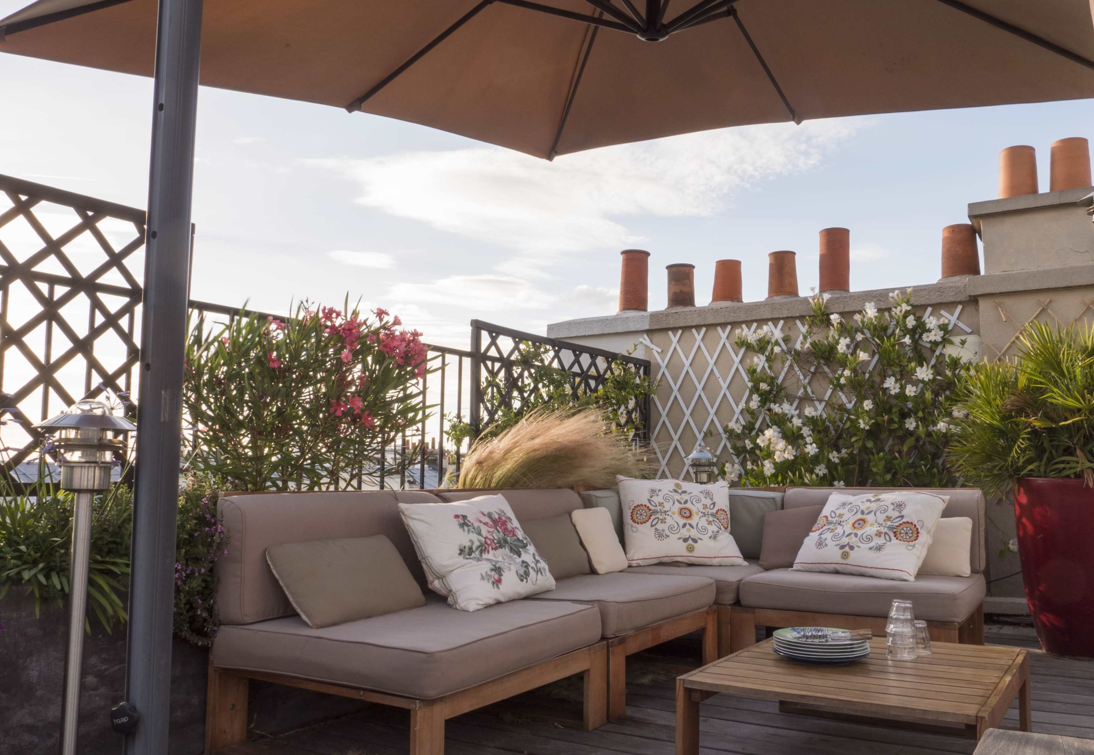A rooftop terrace with a beige sofa set adorned with decorative pillows, a wooden coffee table, and potted plants under a large umbrella.