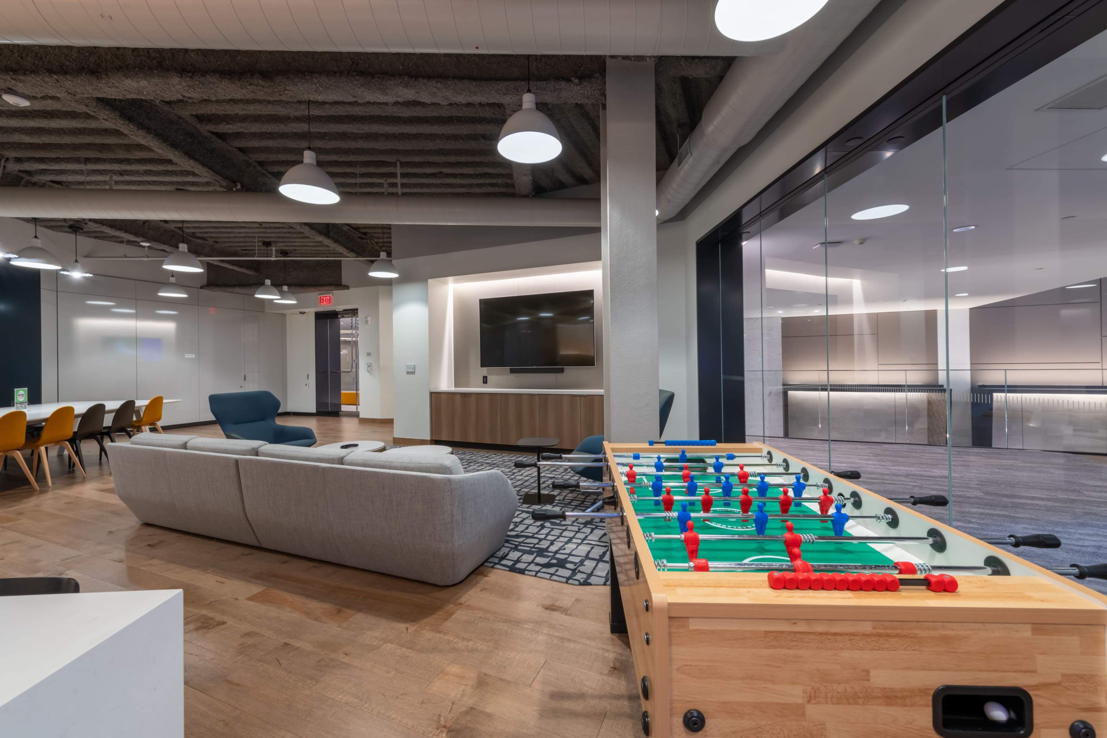 A modern lounge area featuring a foosball table, a gray sofa, and large windows overlooking another space.