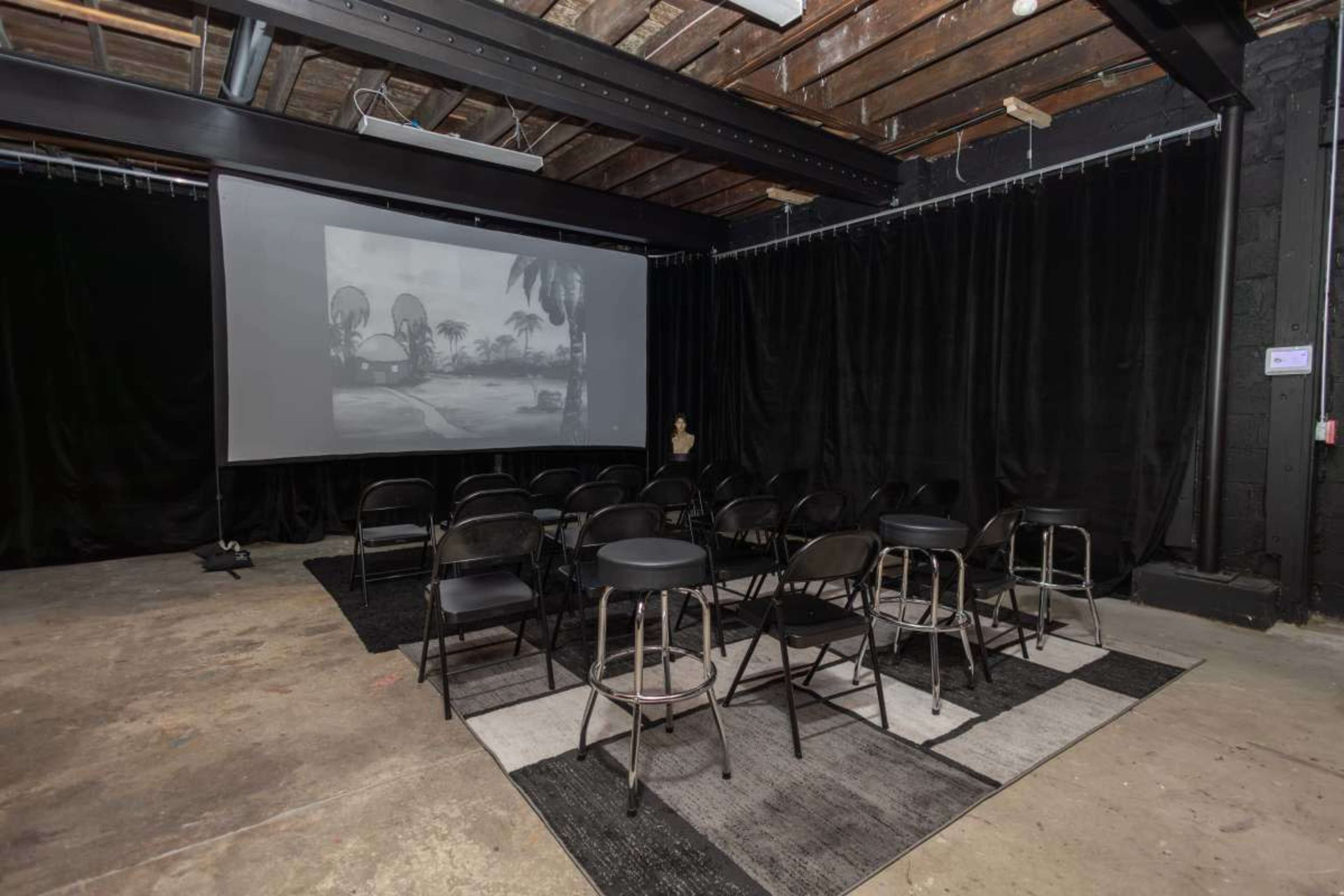 The image shows an indoor space with several black chairs arranged facing a large screen displaying a black-and-white image, surrounded by dark curtains and an unfinished ceiling.