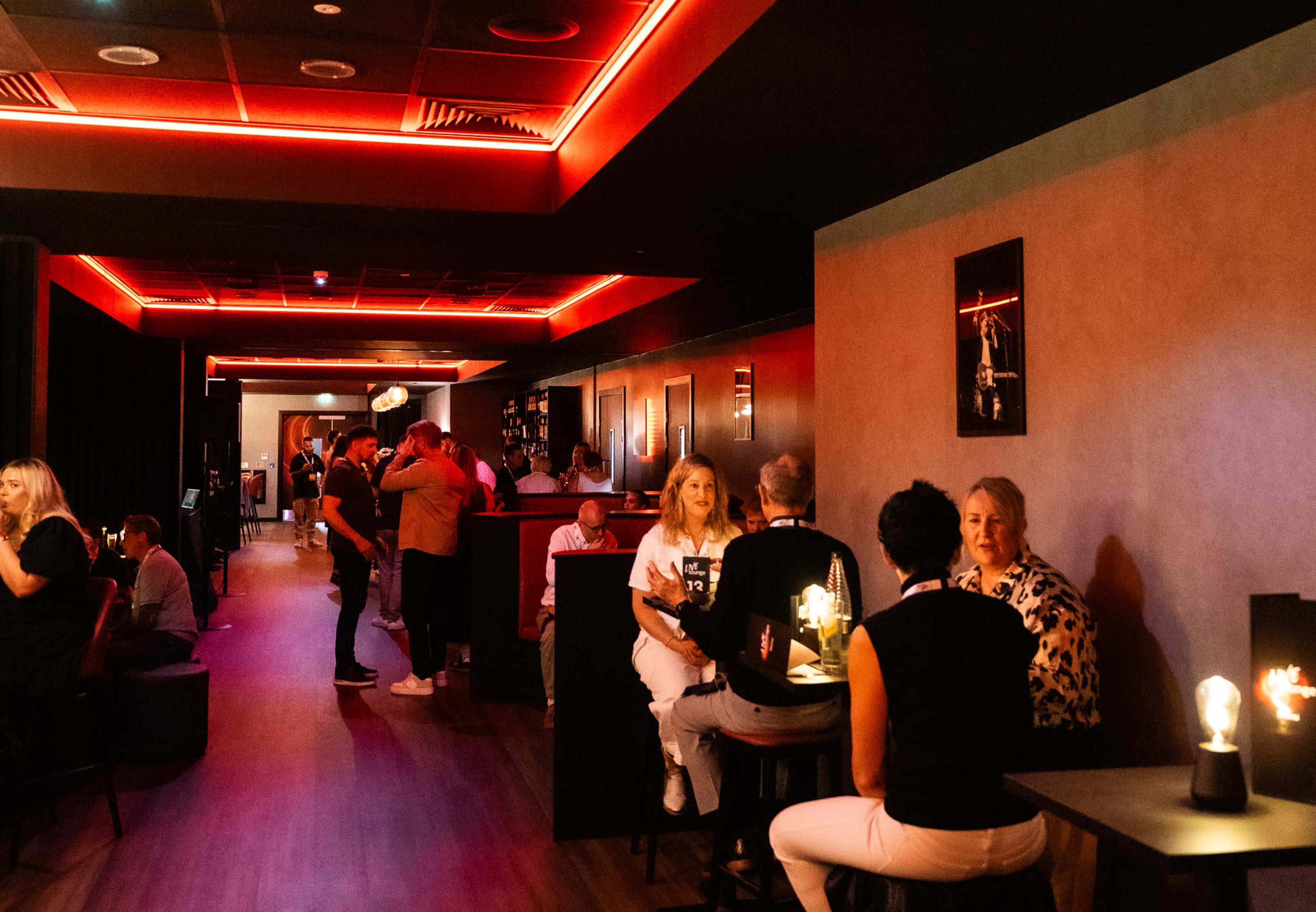 The image shows a dimly lit bar area with low seating, illuminated by red ambient lights, and people socializing at tables and the bar.
