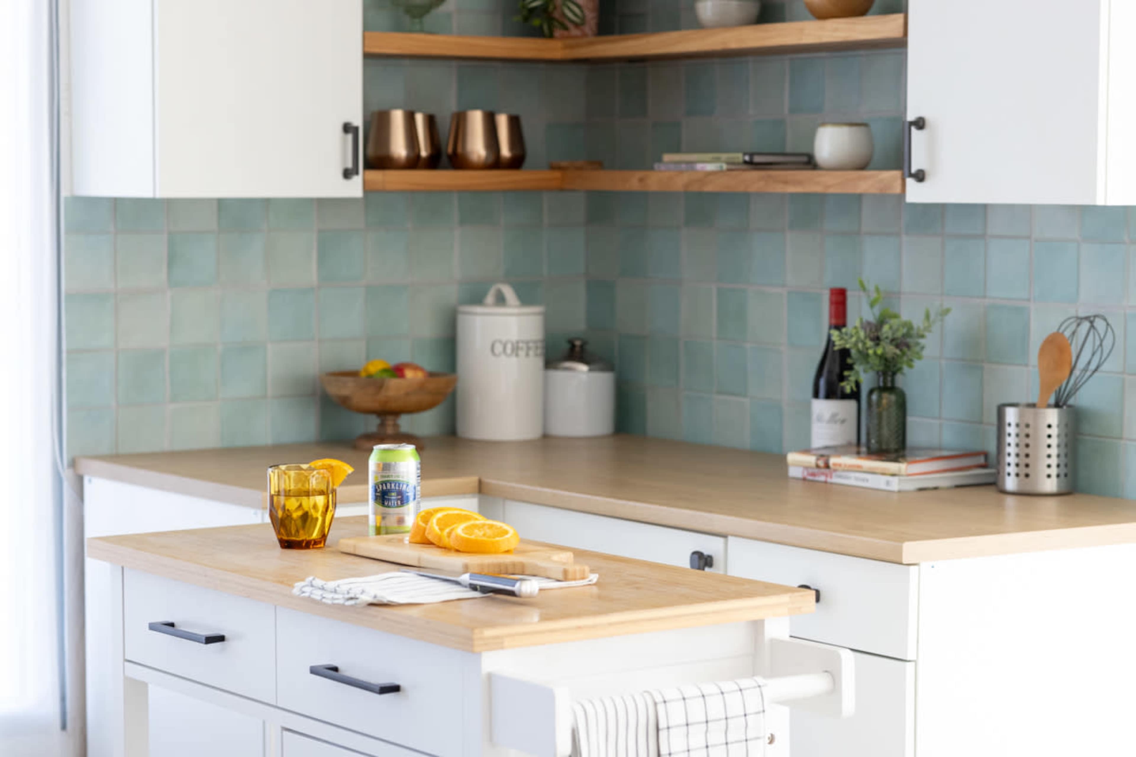 A modern kitchen features a wooden countertop with sliced oranges, two glasses, and a can, along with light blue tiled walls and open shelving displaying various items.