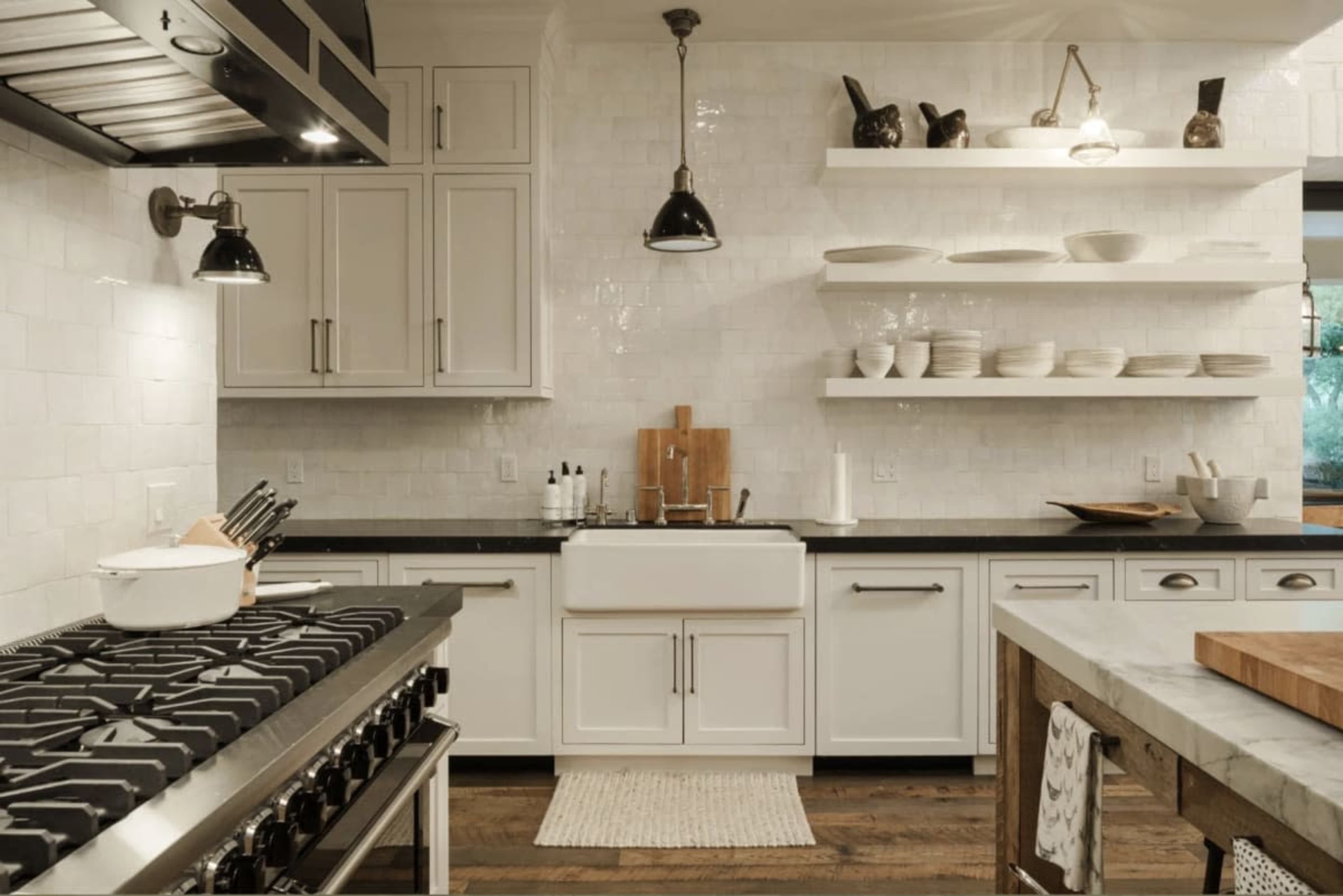 The image shows a modern kitchen featuring white cabinetry, a farmhouse sink, open shelving with dinnerware, and a gas stove.