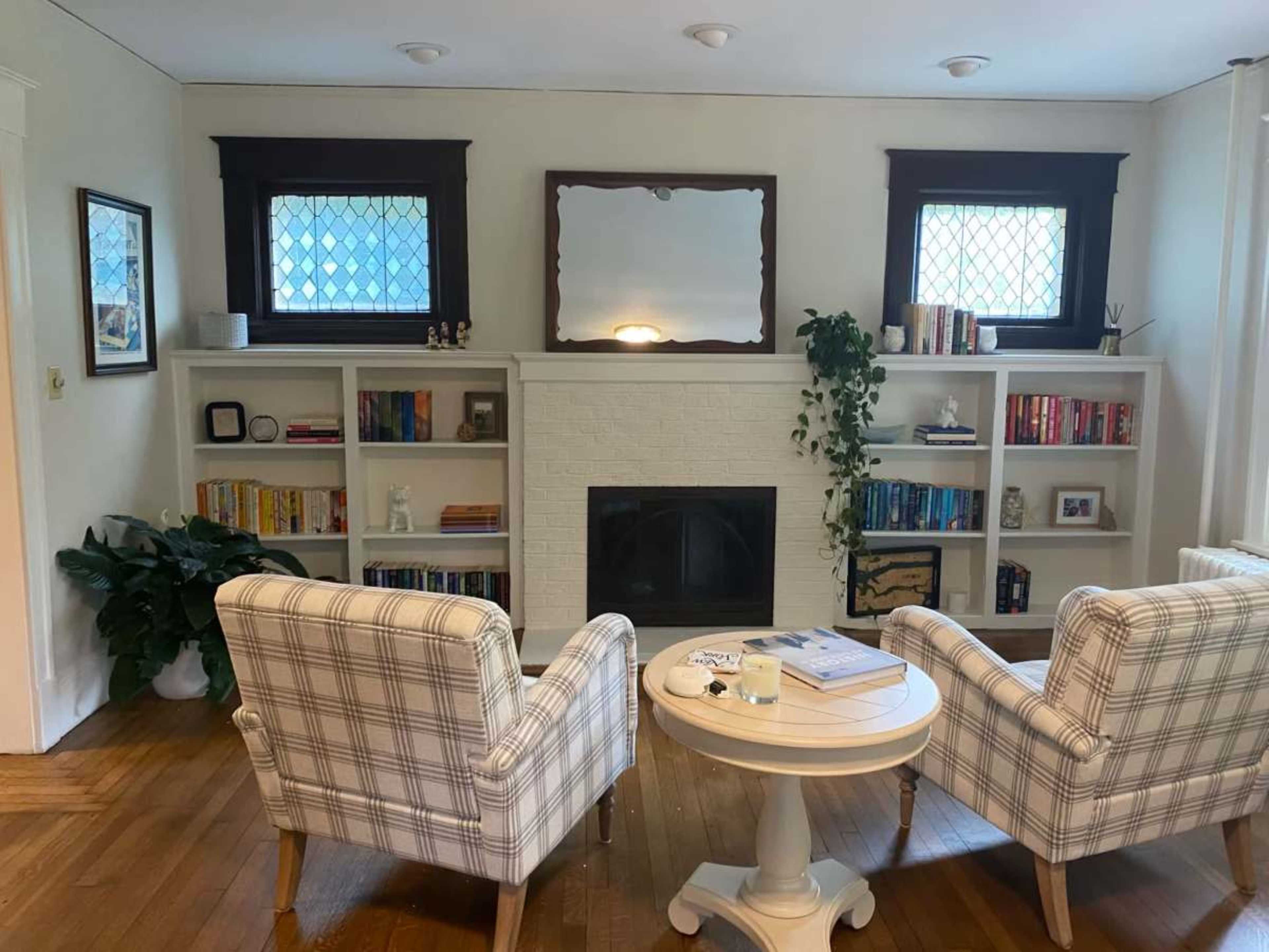 The image shows a cozy living room with two plaid armchairs facing a white fireplace, flanked by built-in bookshelves filled with books and decorative items.
