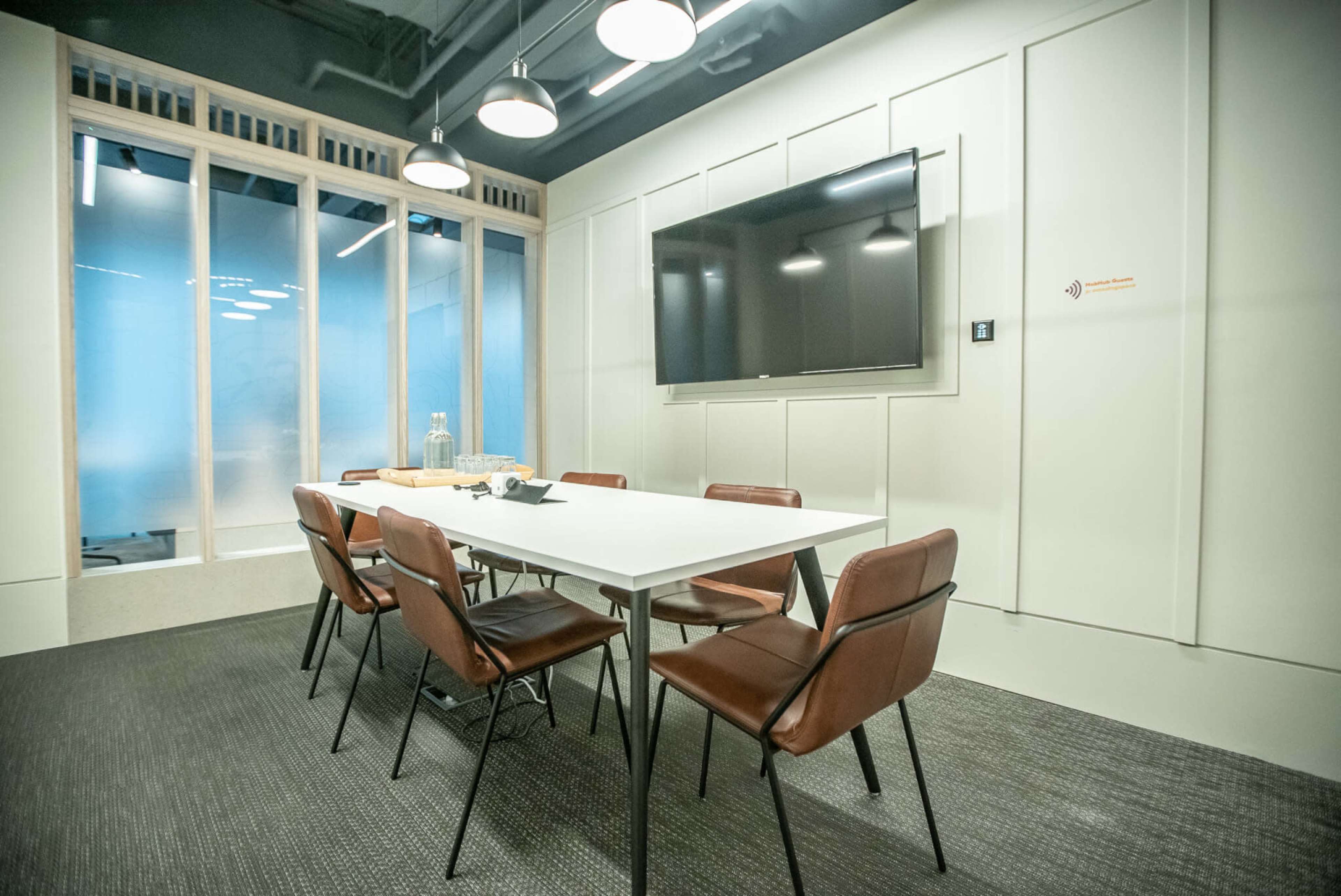 The image shows a modern meeting room with a large table, six chairs, a television mounted on the wall, and glass windows.
