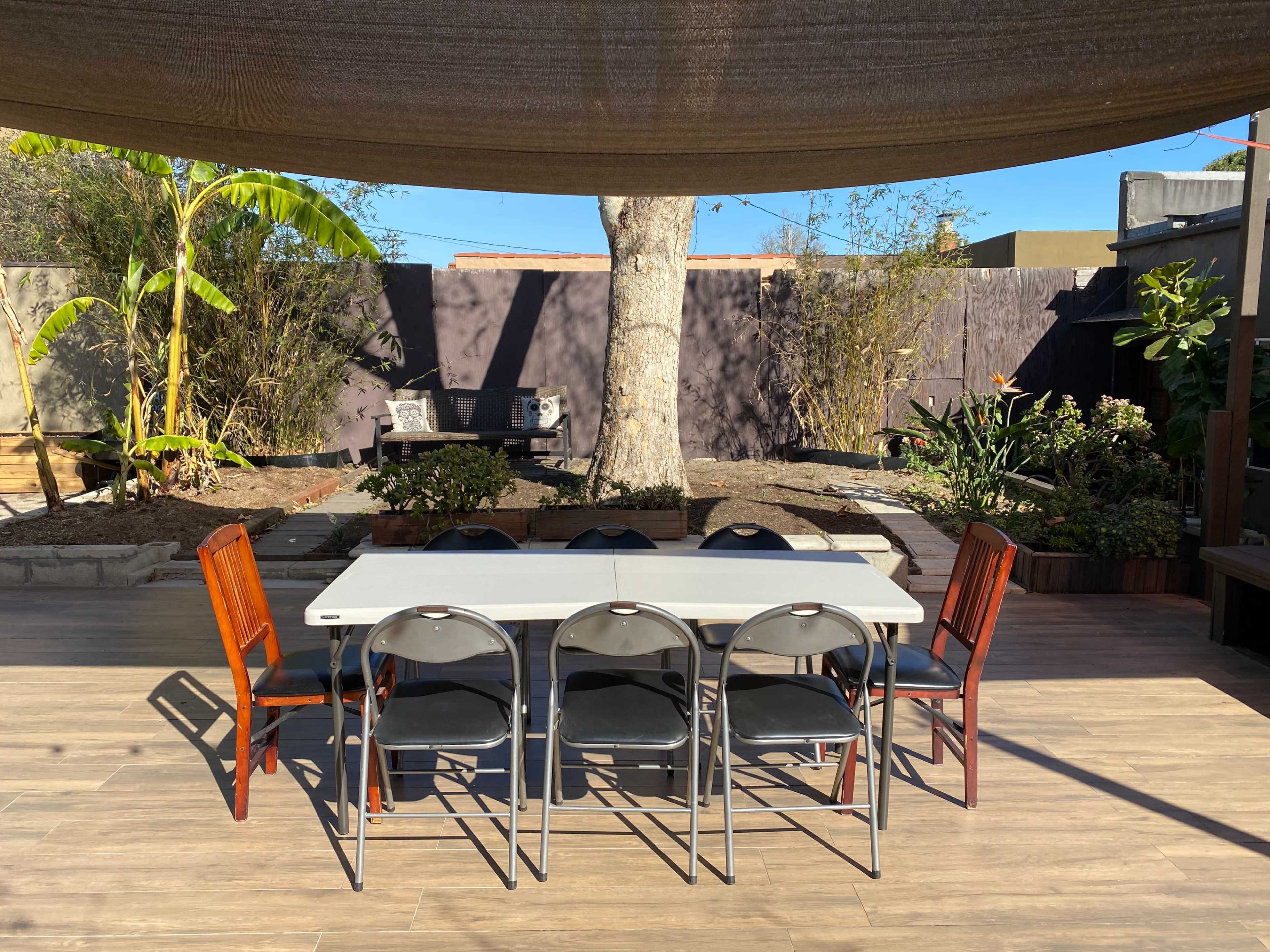 A rectangular table surrounded by six chairs is set in a backyard with a large tree and landscaped greenery in the background.