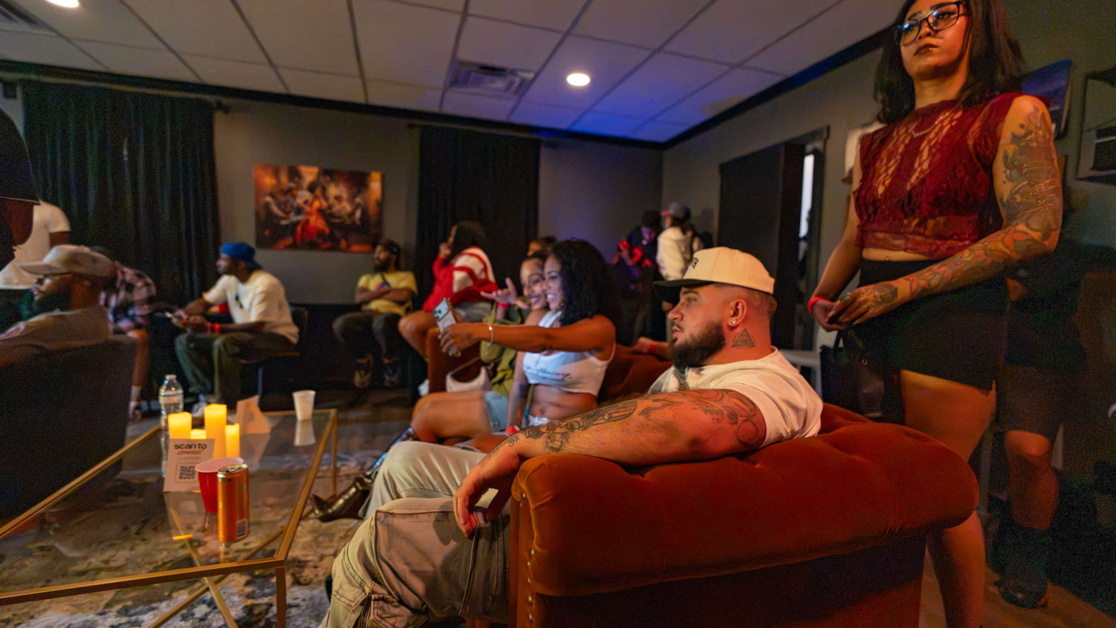 A group of people socializes in a dimly lit lounge, with a mix of seated and standing individuals, while a table displays drinks and candles.
