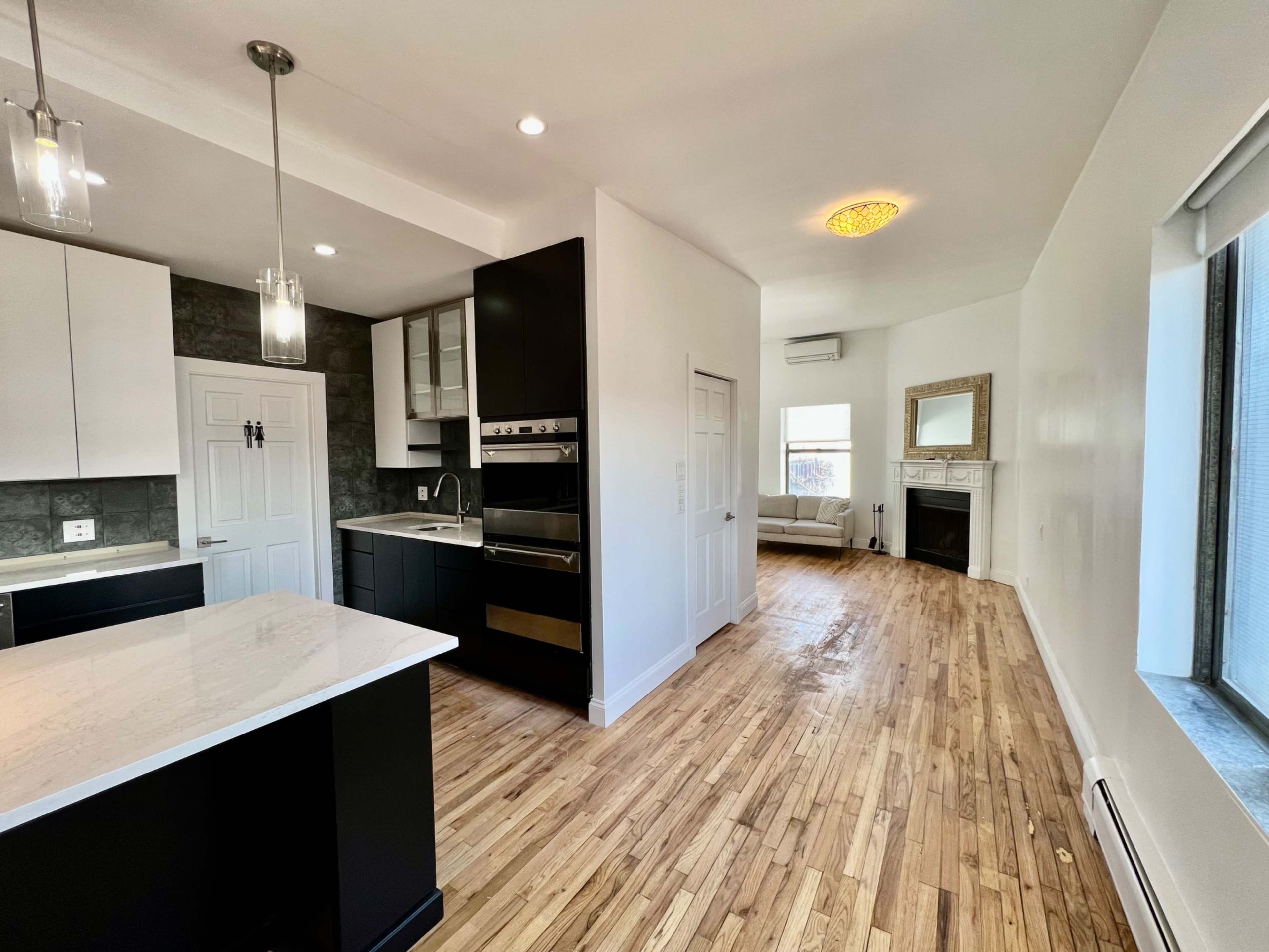 A modern kitchen with black and white cabinetry, a marble countertop, and an adjacent living area featuring a fireplace and a large window.