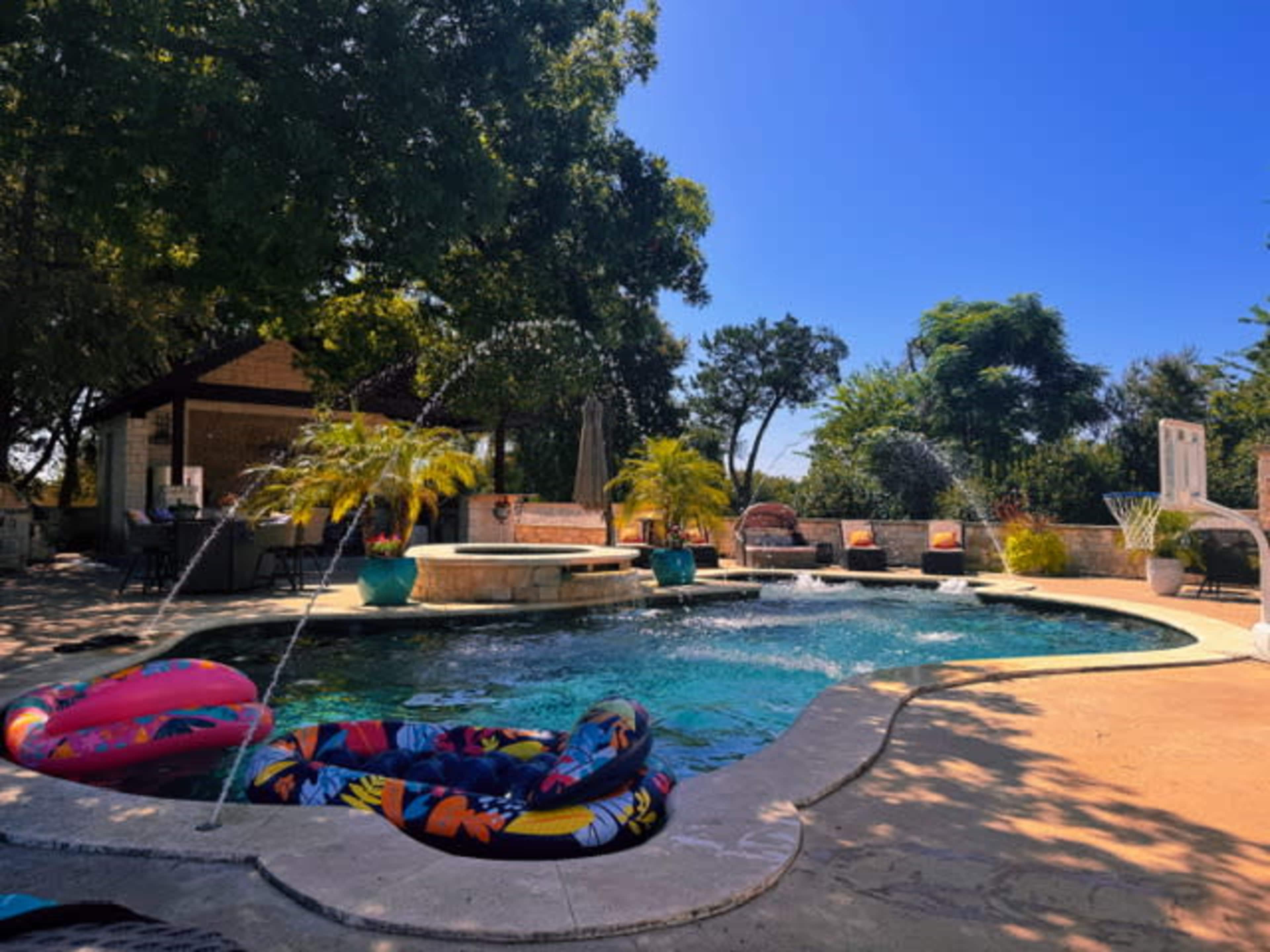 A colorful pool area features lounge chairs, potted plants, and inflatable floats, surrounded by trees and a clear blue sky.