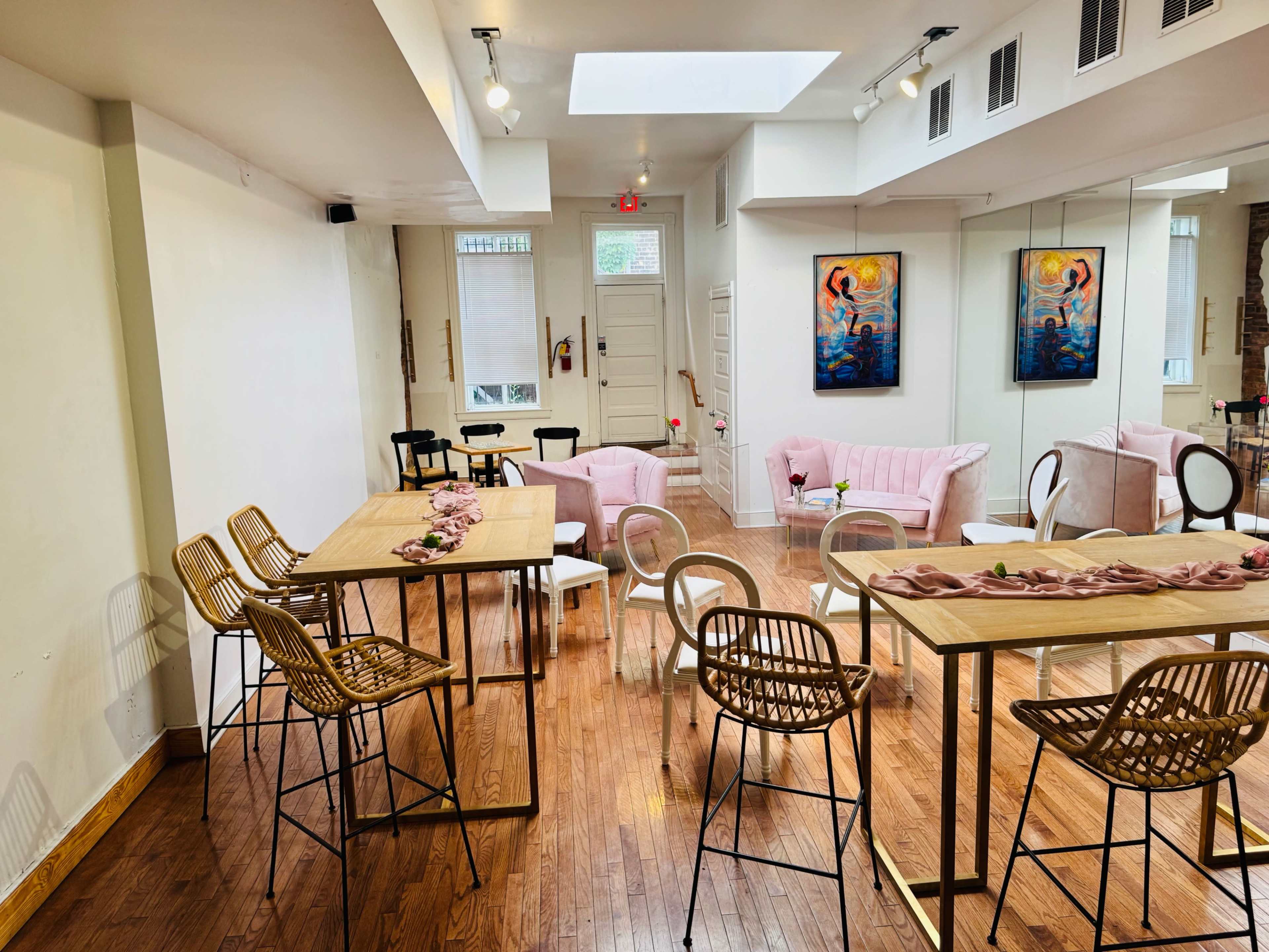 The image shows a brightly lit, modern interior space with wooden floors featuring various seating arrangements, including pink chairs and bar-style tables.