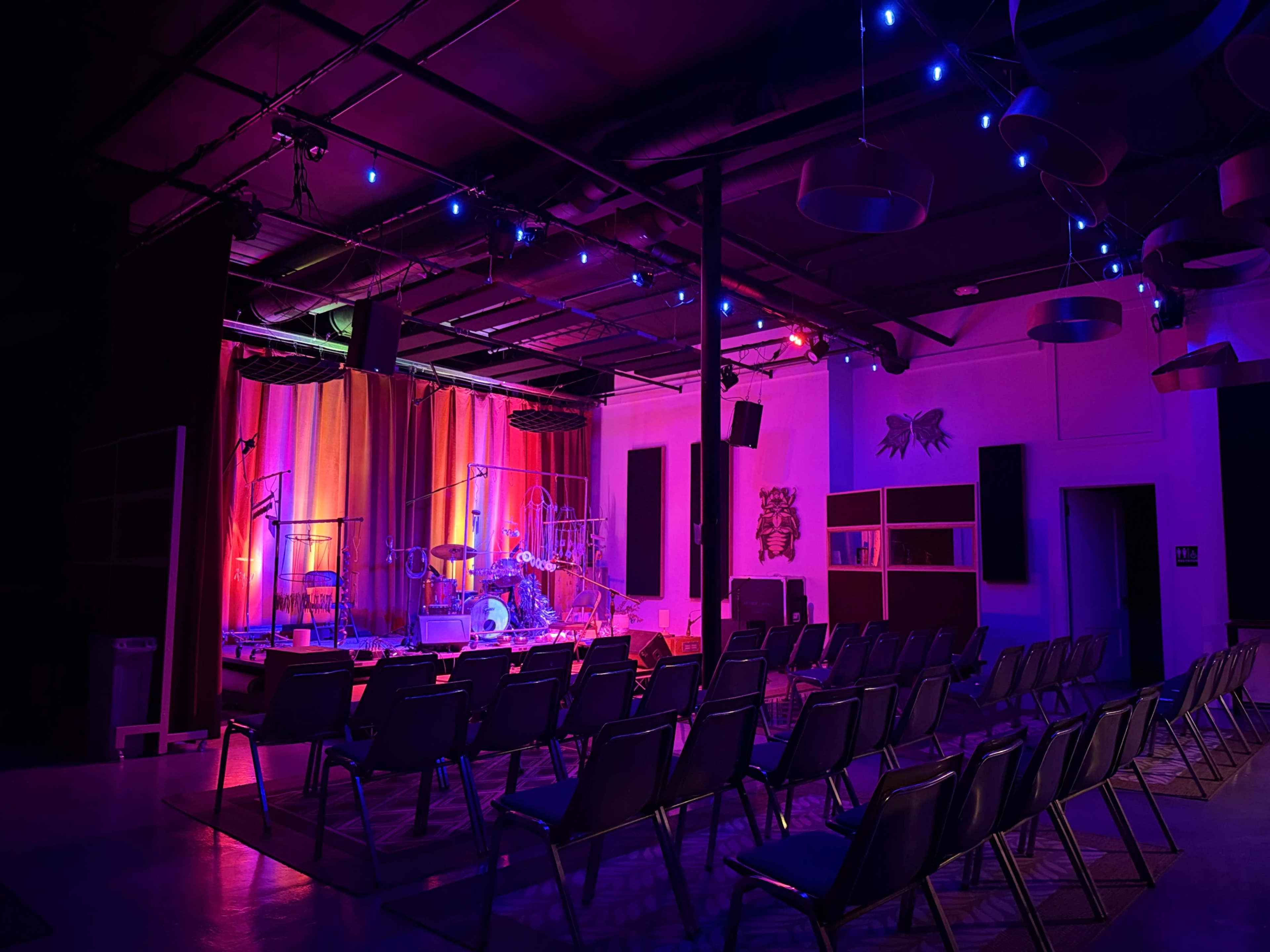 A small performance space features rows of empty chairs facing a brightly lit stage with musical instruments and colorful lighting.