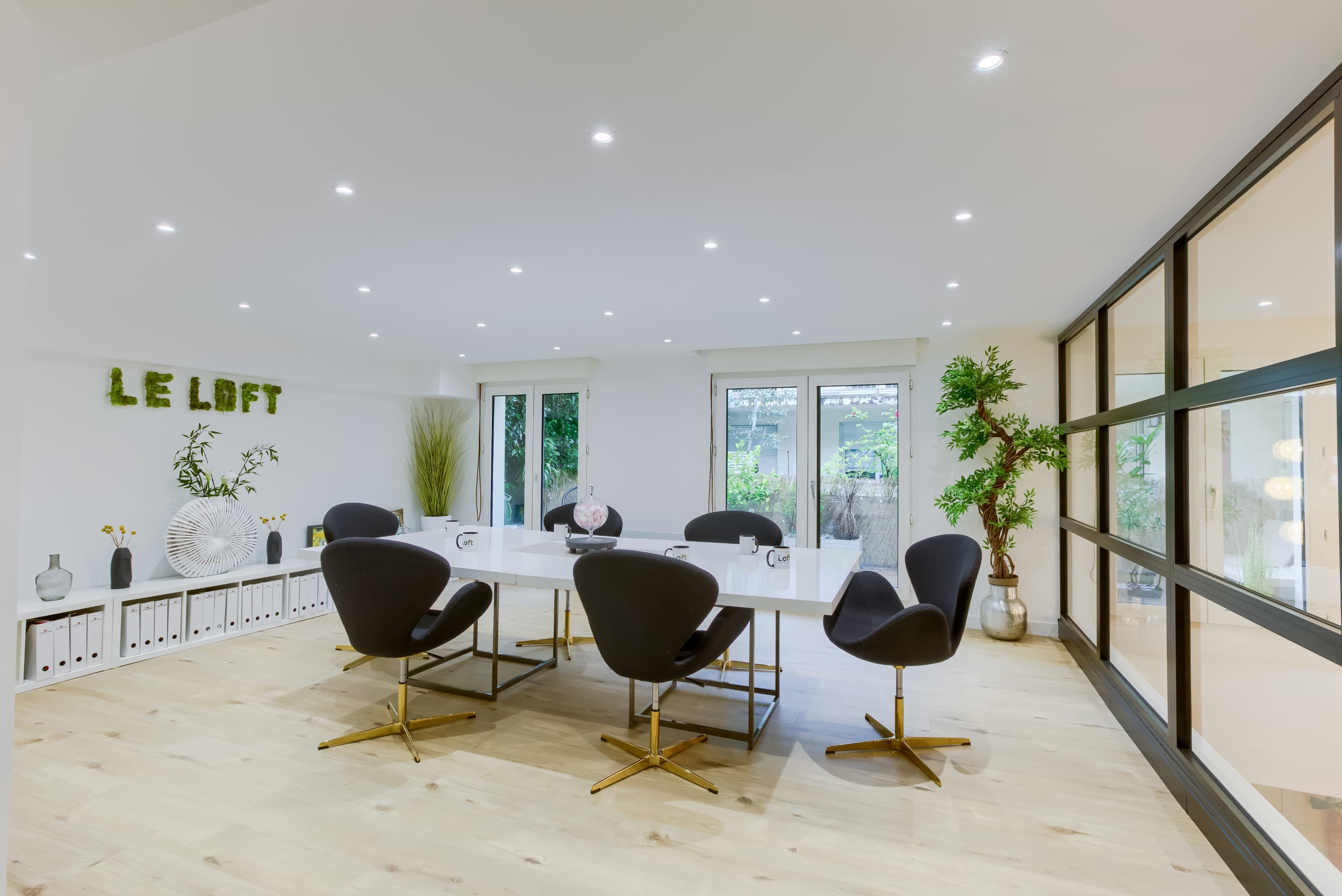 A modern conference room features a large white table surrounded by black chairs, with glass doors leading to an outdoor area and greenery in the decor.