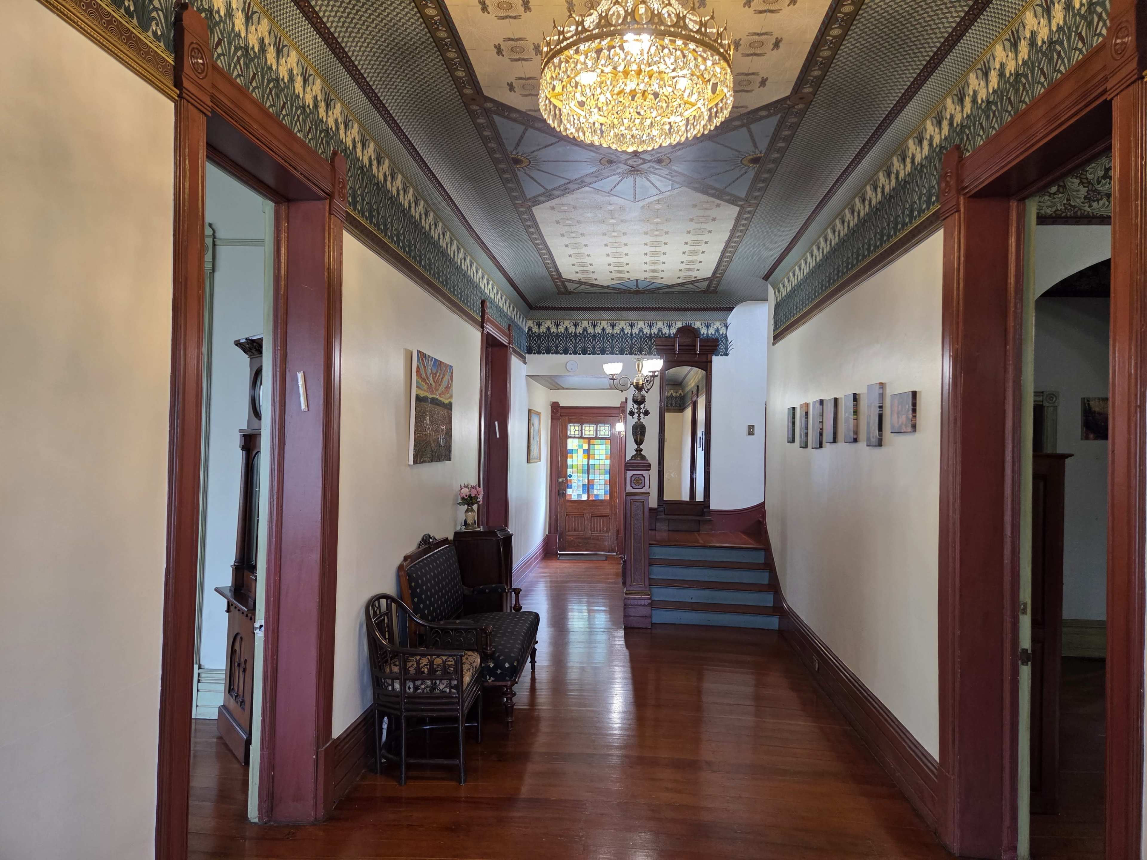 The image shows a long hallway with wooden flooring, a decorative ceiling, and a chandelier, flanked by doorways leading to adjacent rooms.