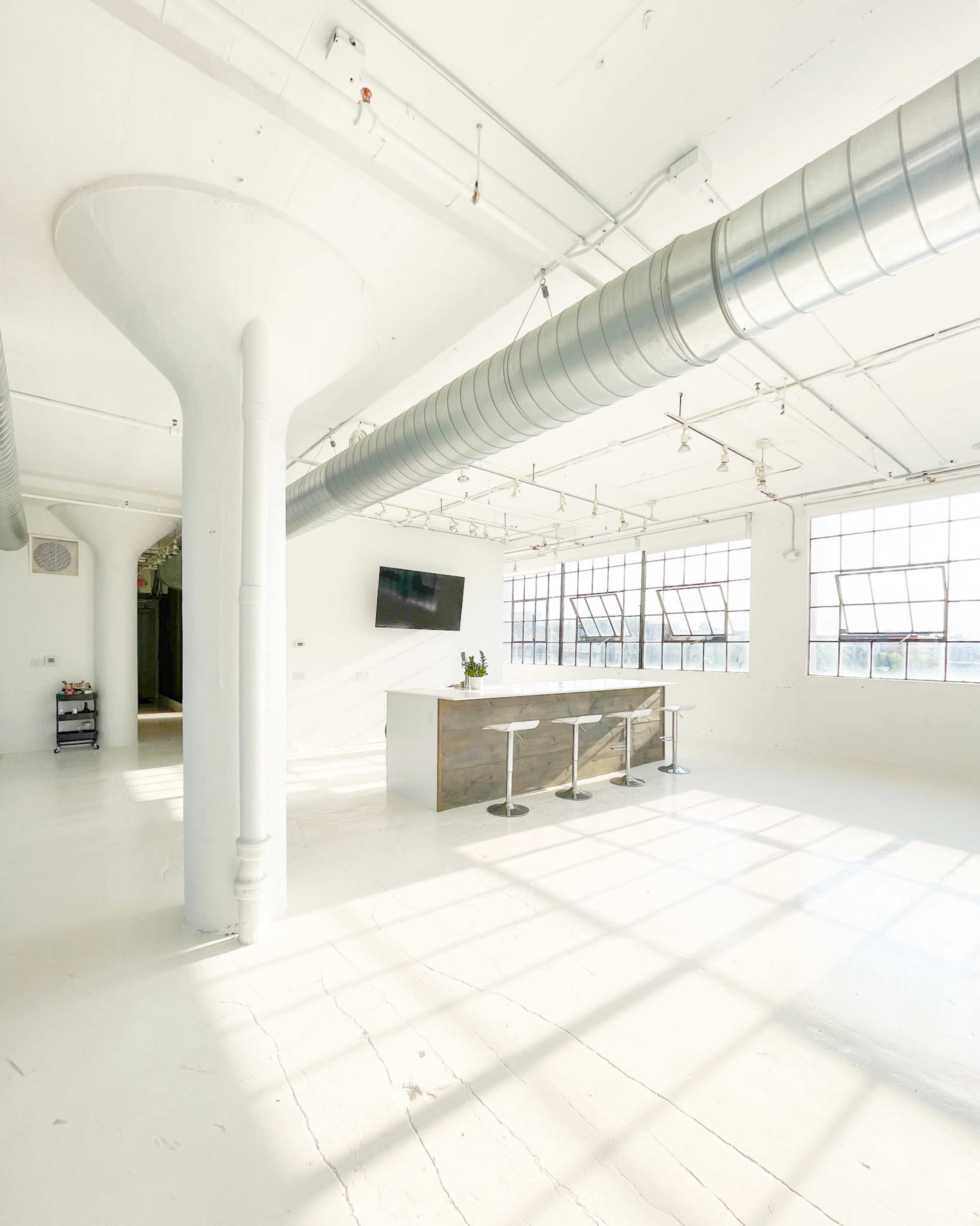 The image shows a bright, open space with white walls and floors, featuring a bar with three stools and large windows allowing natural light to fill the area.