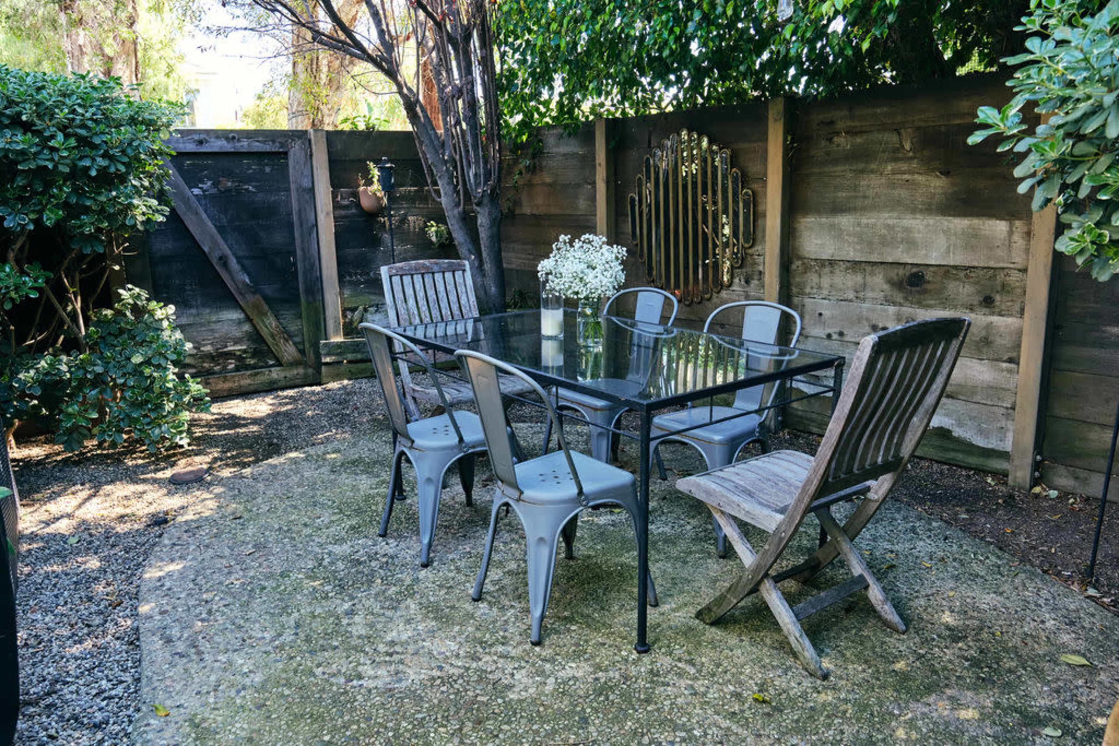 The image shows a small outdoor seating area with a glass table surrounded by six chairs, set against a wooden fence and greenery.