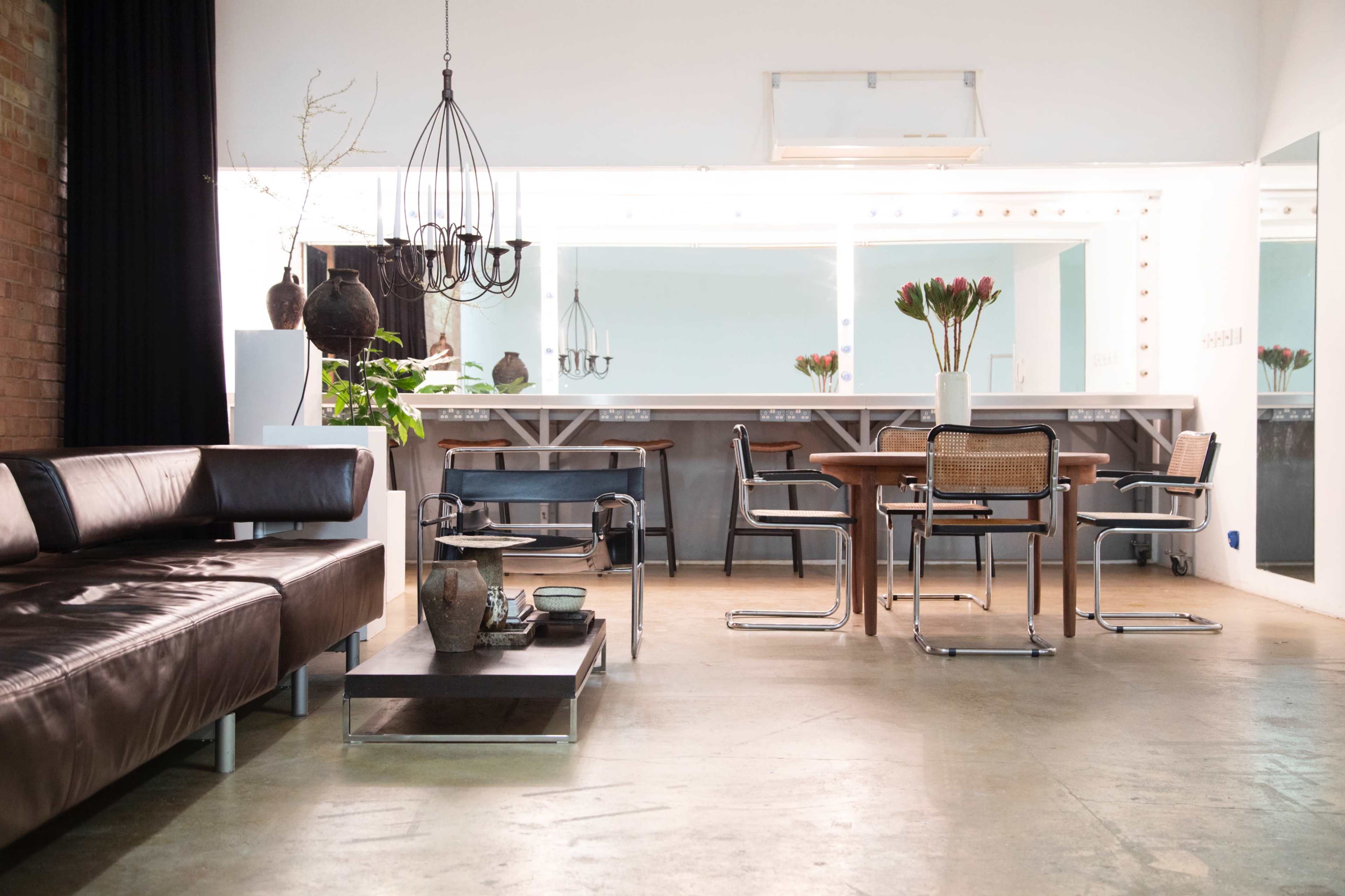 A modern interior space featuring a leather sofa, a dining table with chairs, and a kitchen area with a large mirror and decorative plants.