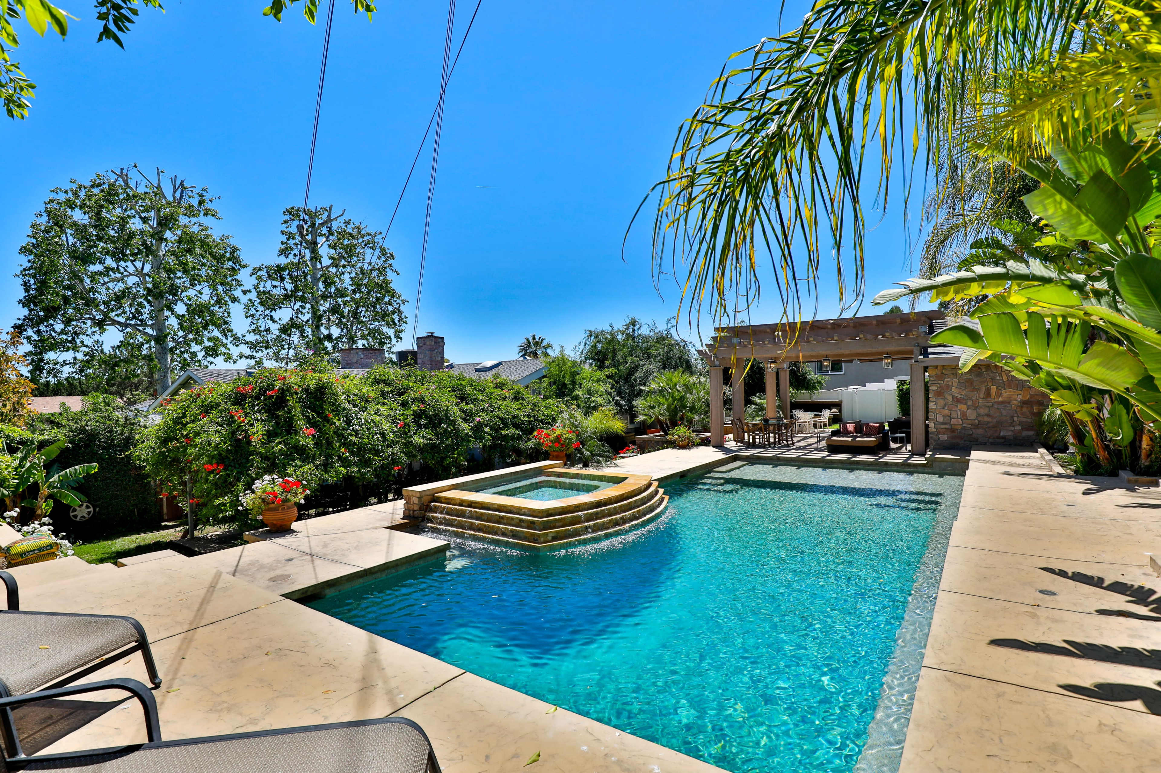 A backyard features a swimming pool with a spa, surrounded by greenery and outdoor furniture under a pergola.