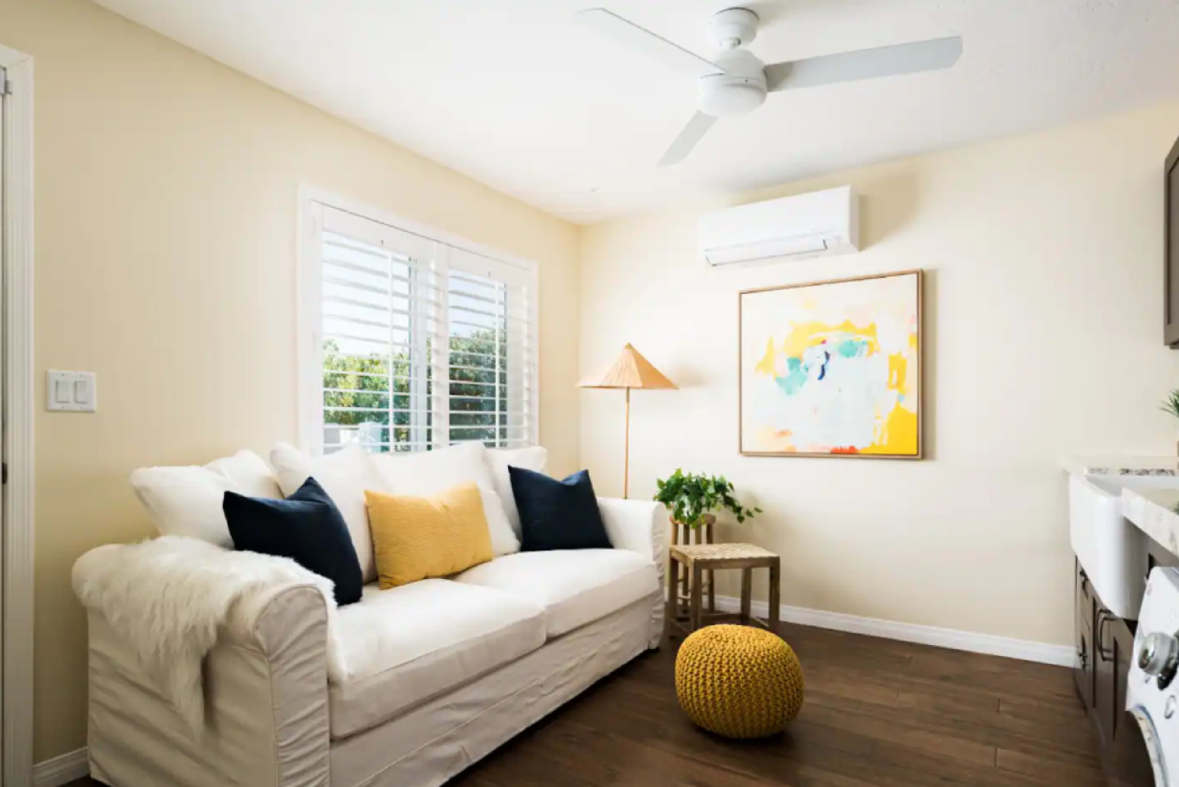 A bright living room features a white sofa with yellow and navy pillows, a wooden side table, a yellow pouf, and a piece of abstract art on the wall.