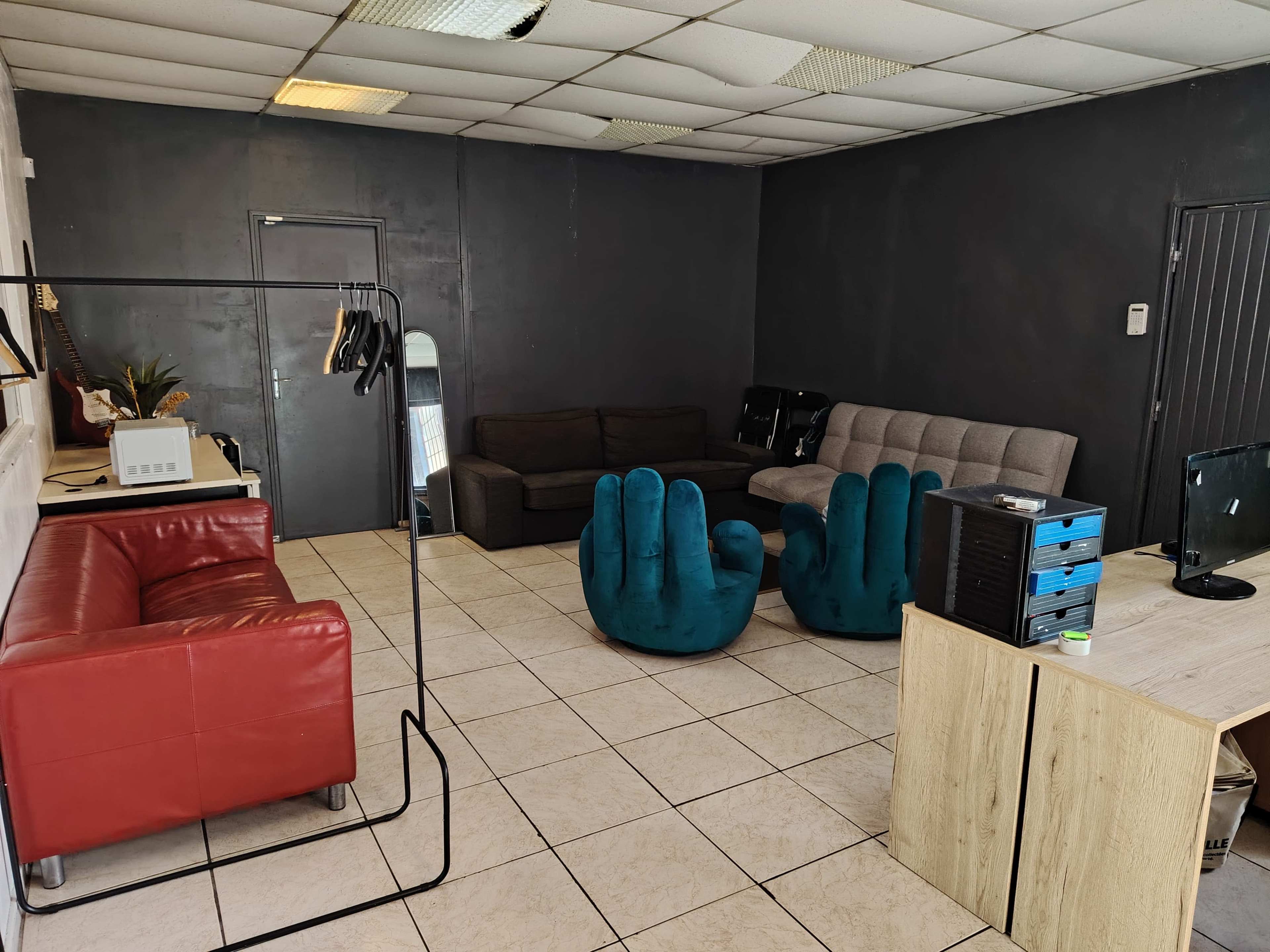 The image shows a sparsely furnished room with various seating options, including a red couch, gray sofas, and blue hand-shaped chairs, along with a desk and a clothing rack.