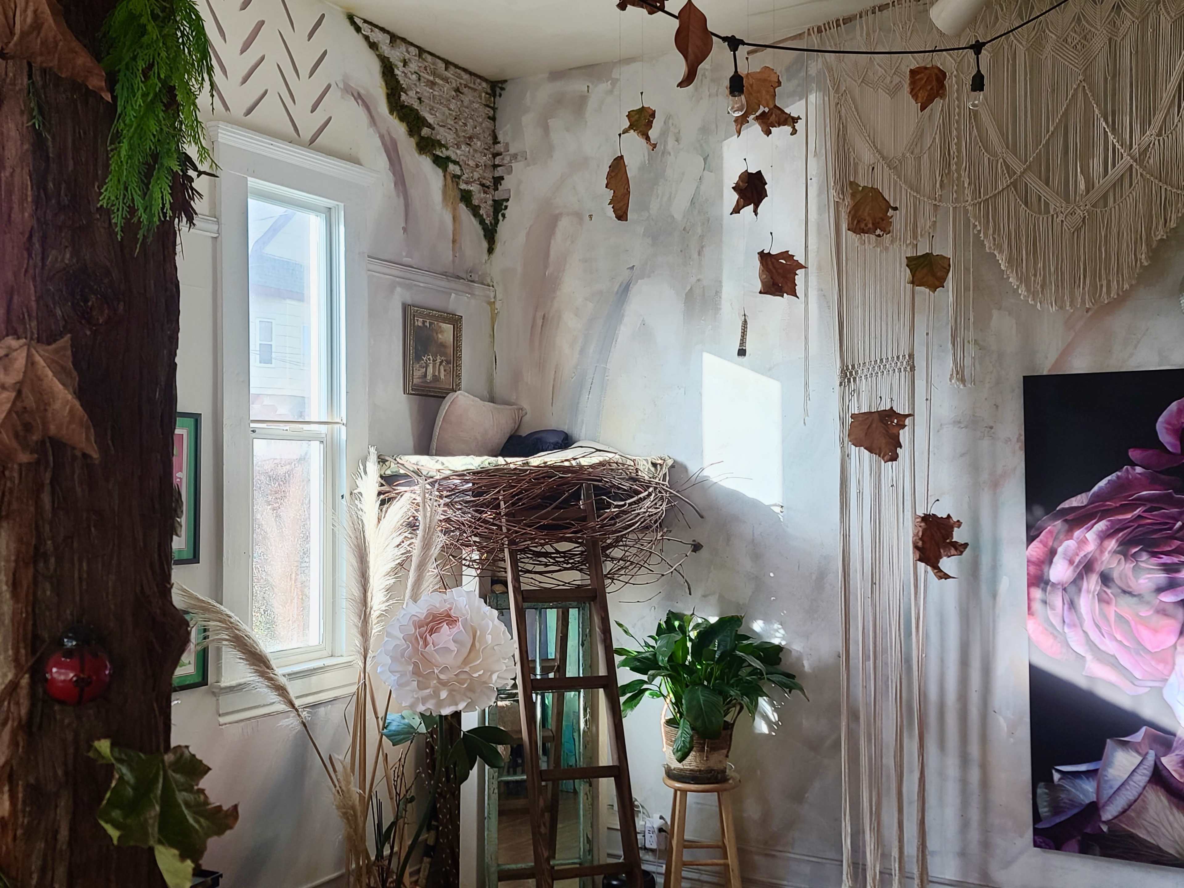 The image shows a room with an artistic mural, a nest-like structure on a wooden stool, plants, and decorative elements hanging from the ceiling.