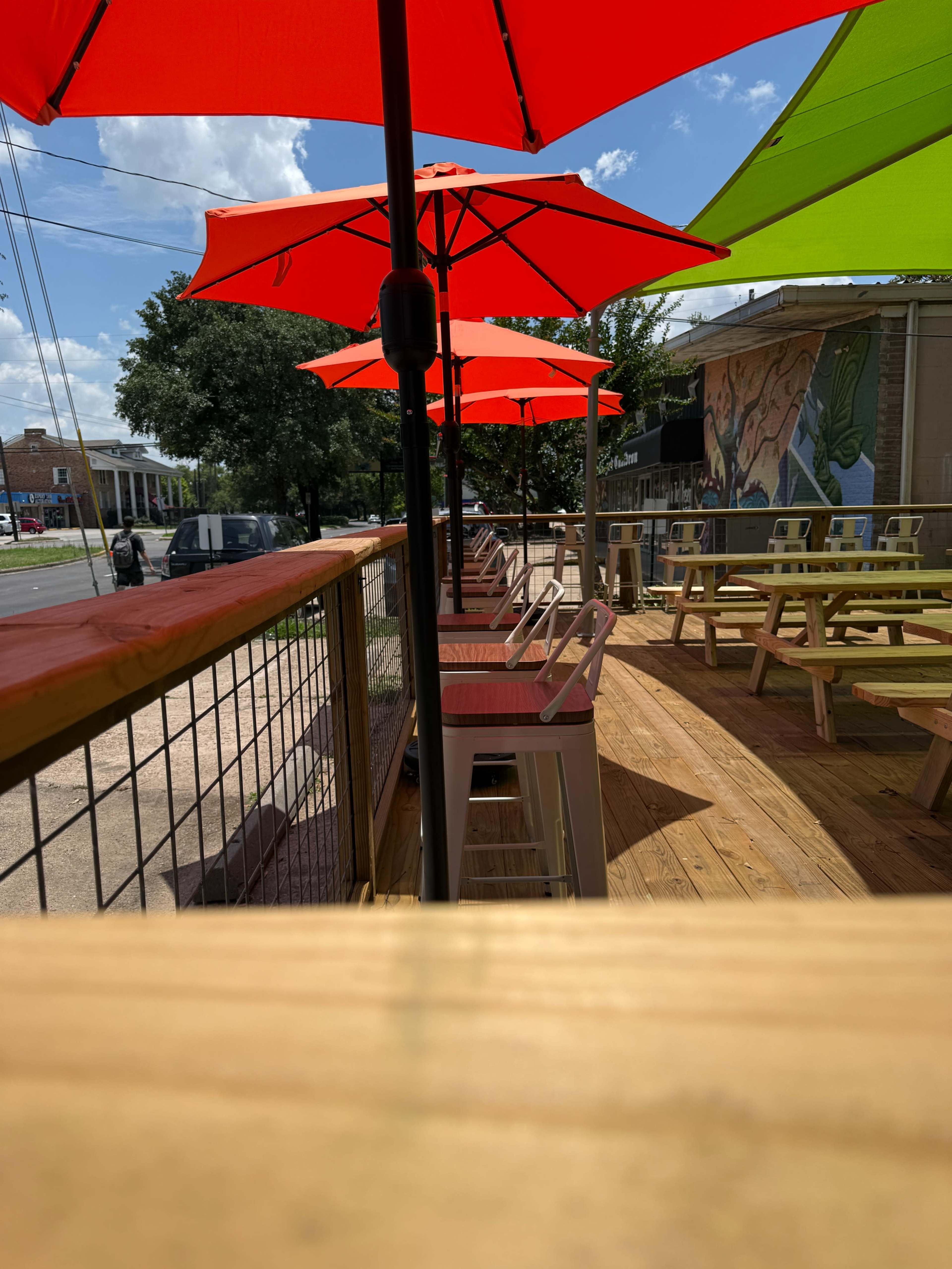 The image shows a wooden outdoor seating area with bright red and green umbrellas on a sunny day.