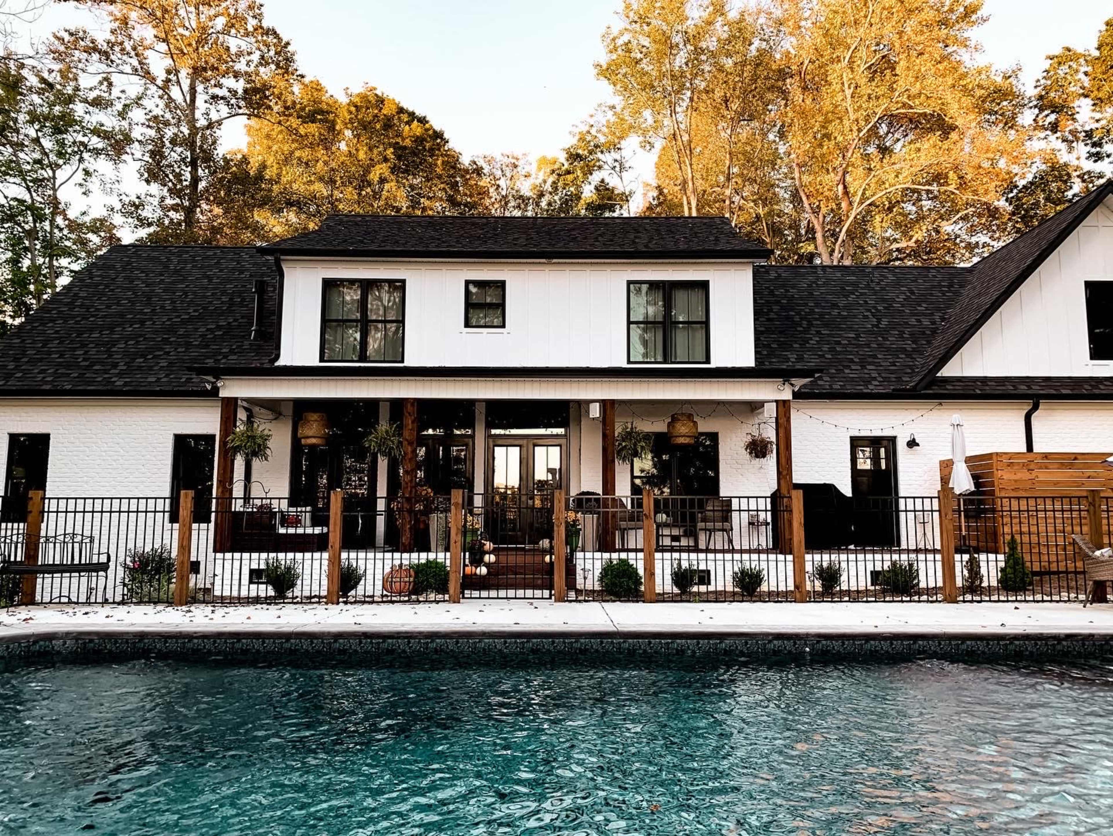 A two-story, modern farmhouse with a black roof and white exterior features a fenced patio area in front of a swimming pool.