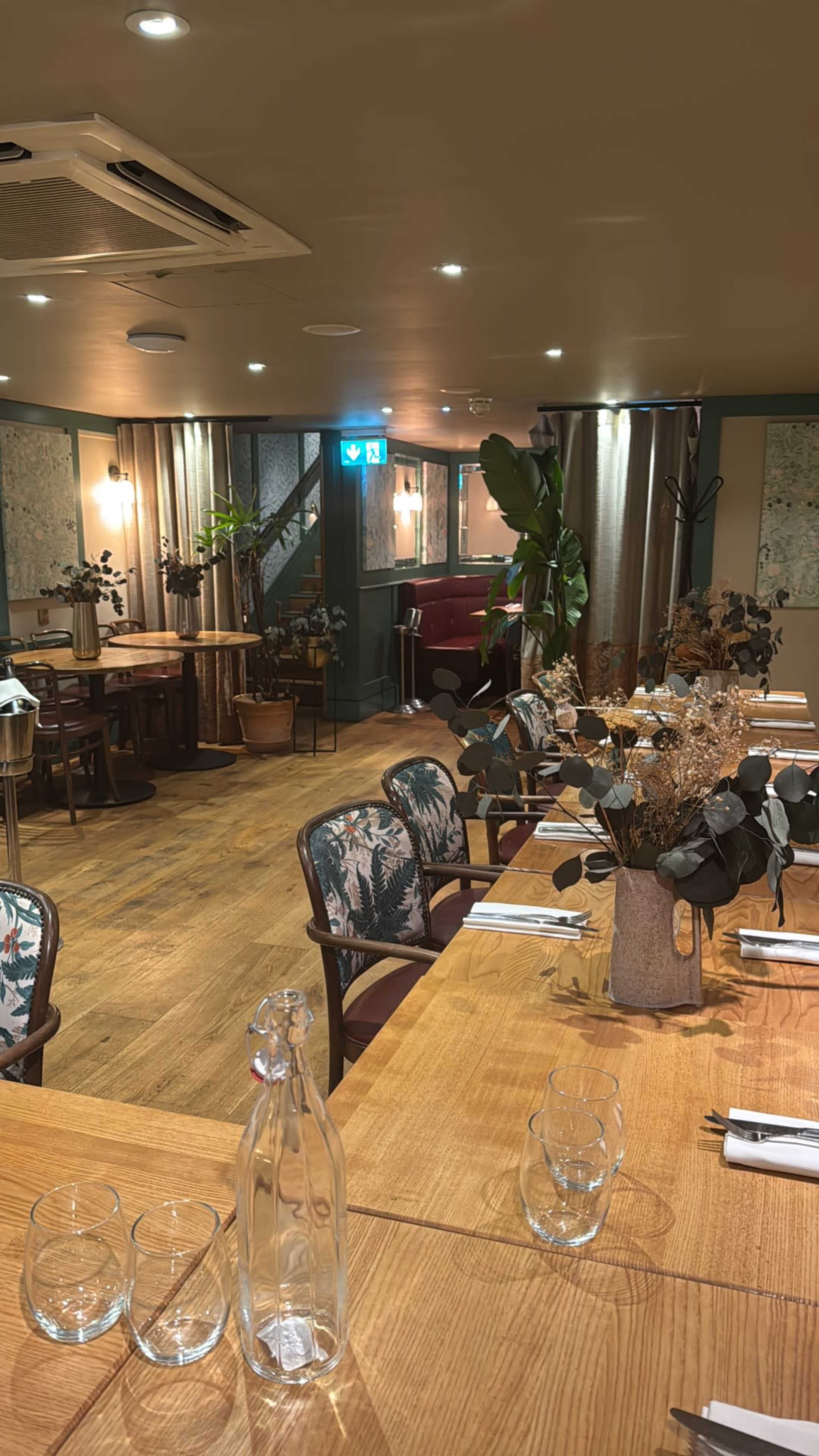 The image shows an elegantly arranged restaurant interior with wooden tables, greenery, and soft lighting.