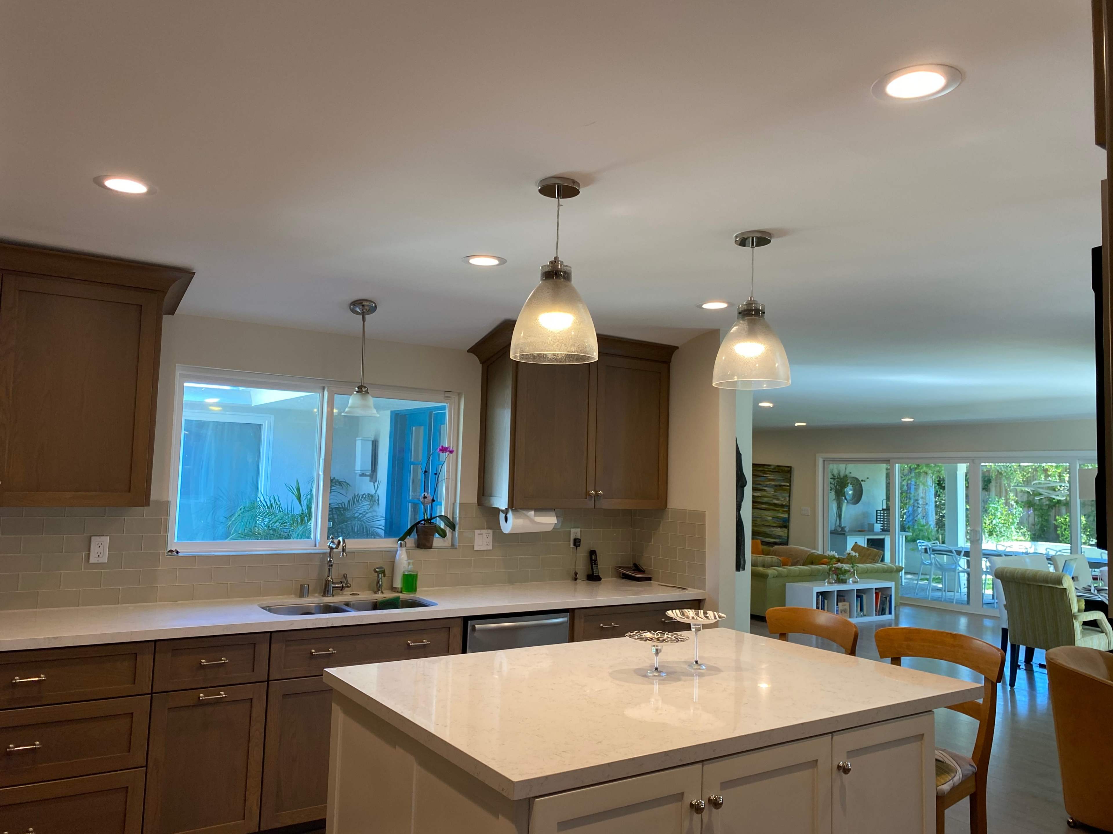 The image shows a modern kitchen with wooden cabinetry, a central island, and pendant lighting, connected to an open dining area featuring large windows.