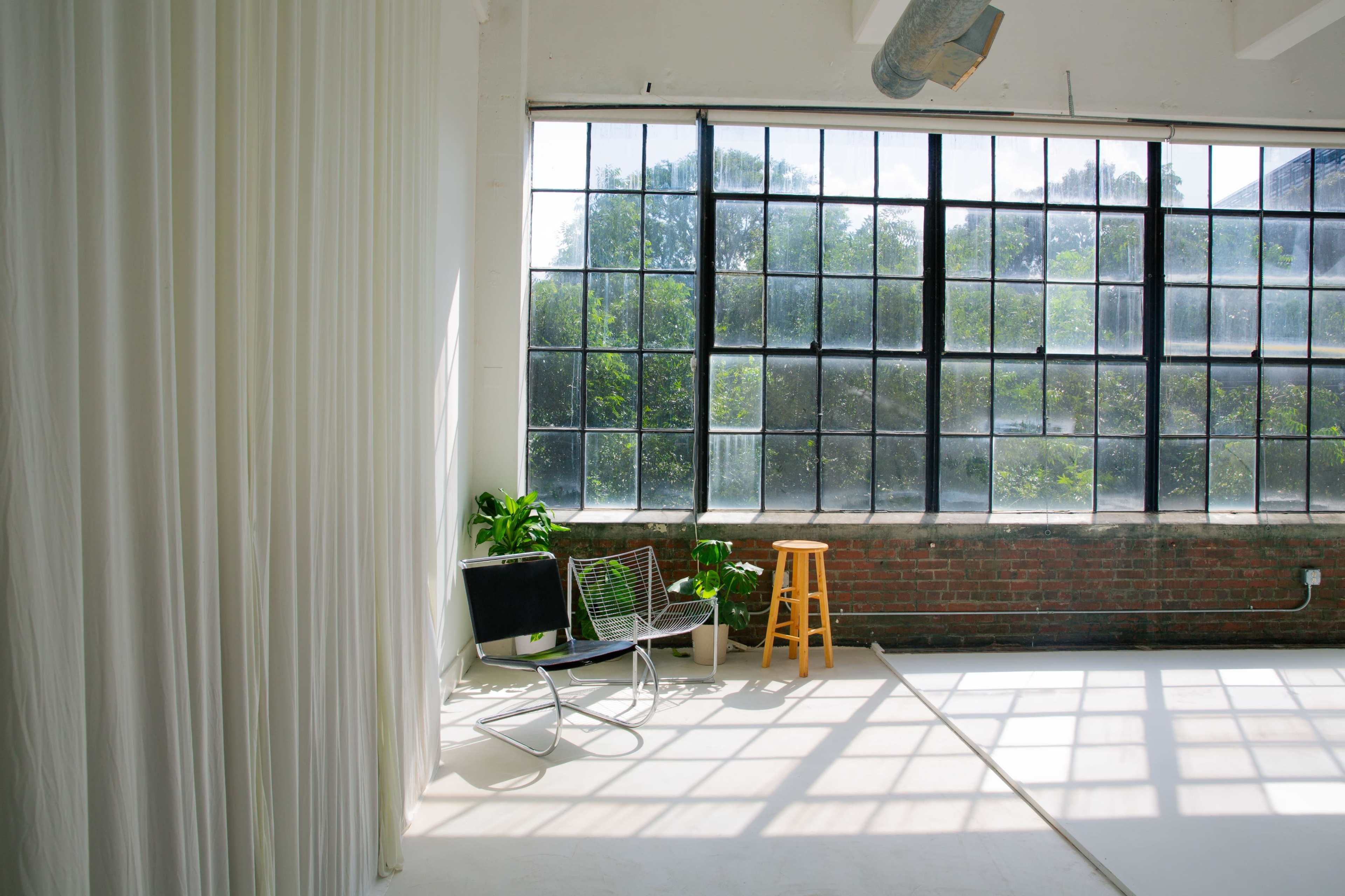 The image shows a bright studio space with large windows, white curtains, a potted plant, a chair, and a wooden stool beside a wooden brick wall.