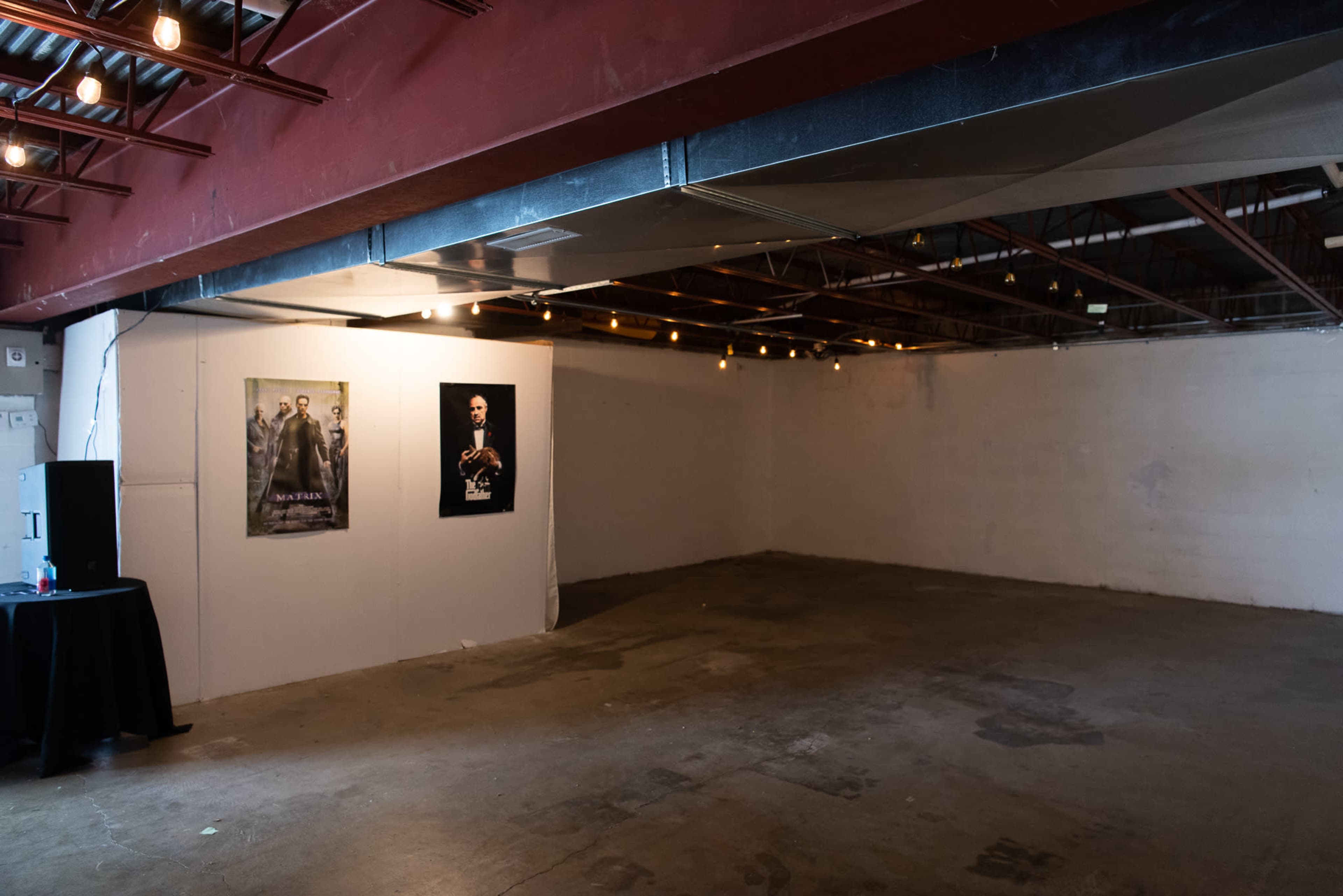 An empty room with bare concrete flooring features two posters on one wall and string lights overhead.