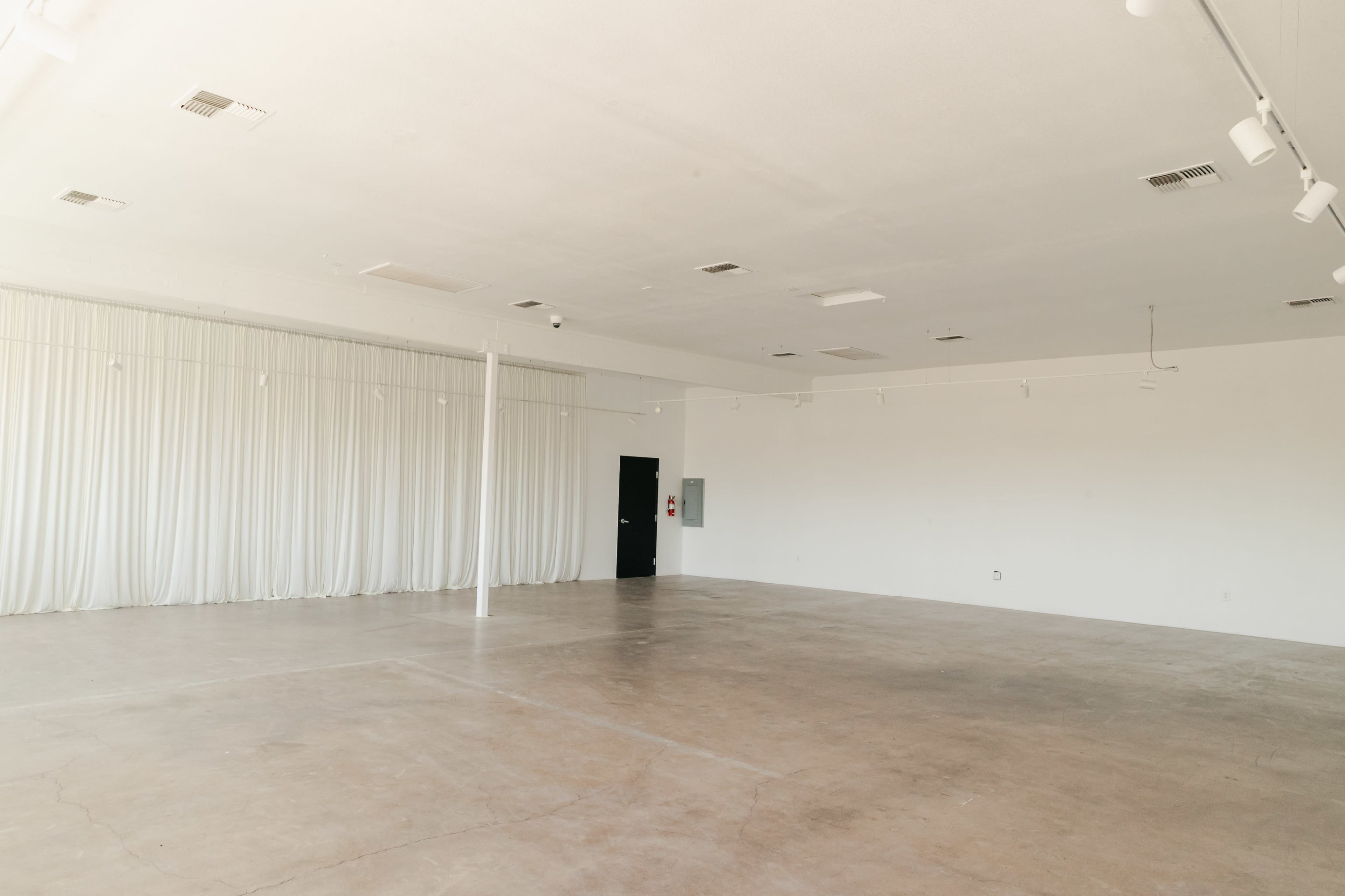 The image shows a spacious, empty room with concrete flooring, white walls, and a large curtain-covered window.