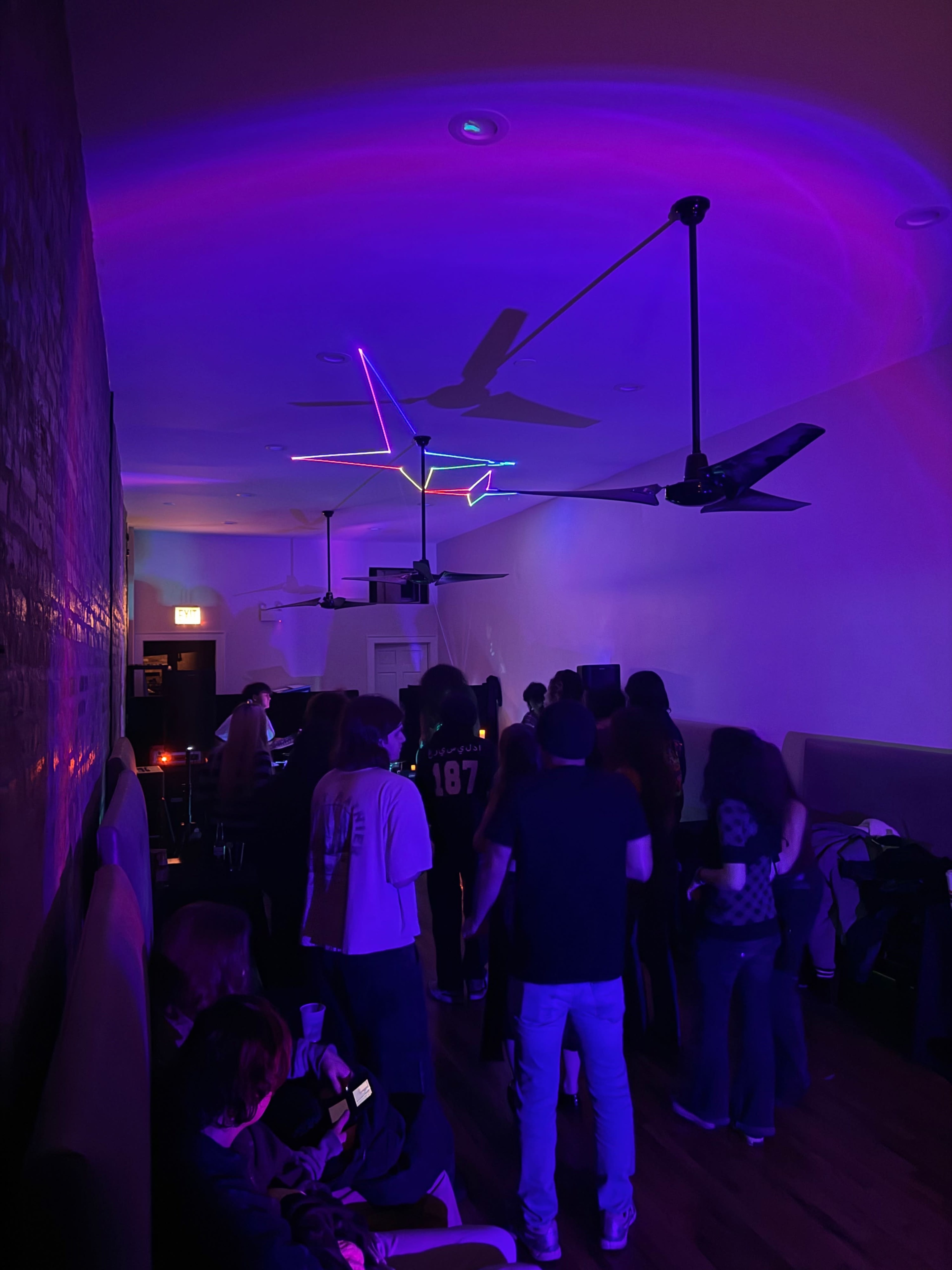 A group of people gathers in a dimly lit room with colorful neon lights and ceiling fans.
