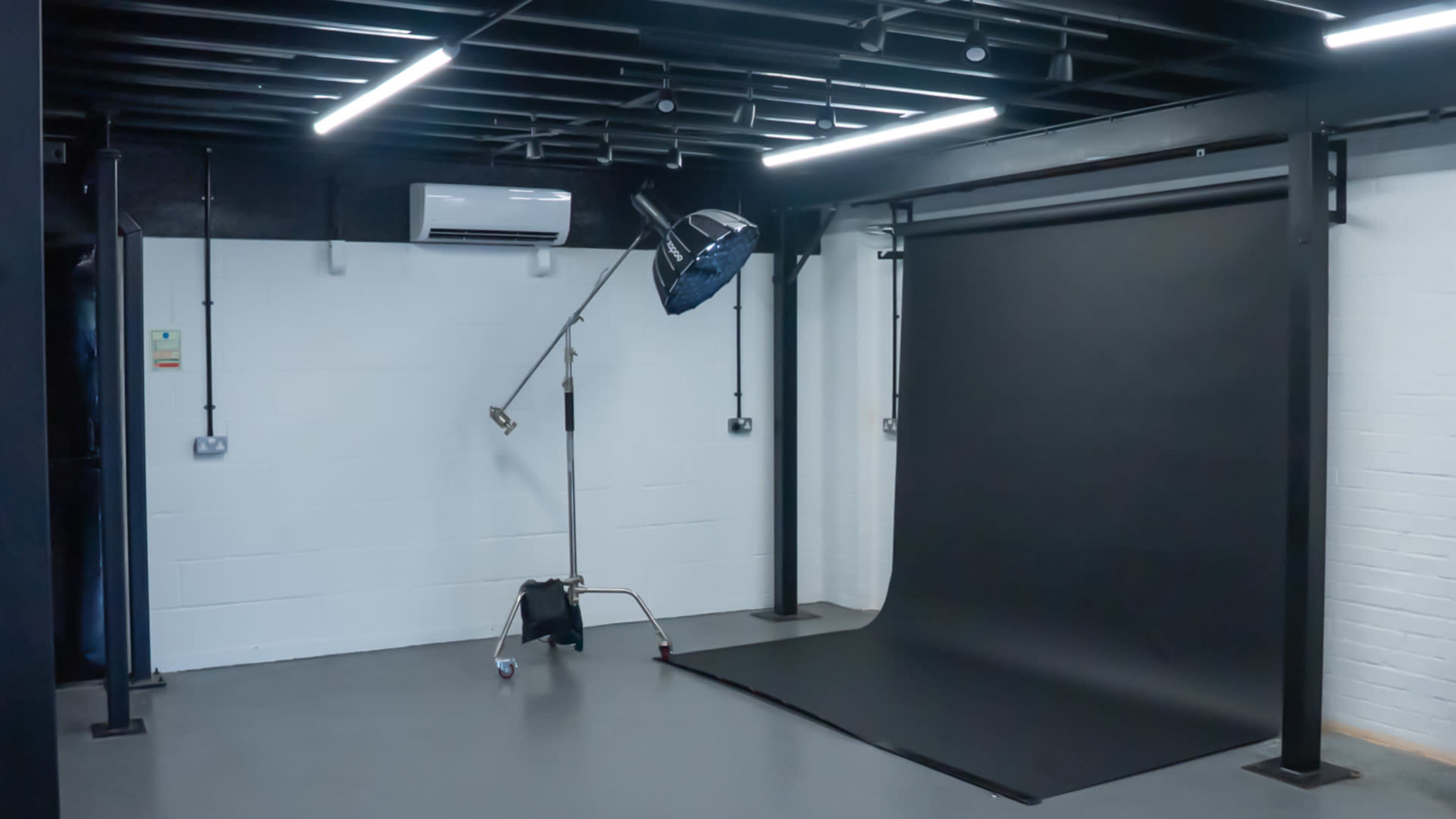 The image shows a photography studio with a black backdrop, a light stand, and ceiling-mounted lights.