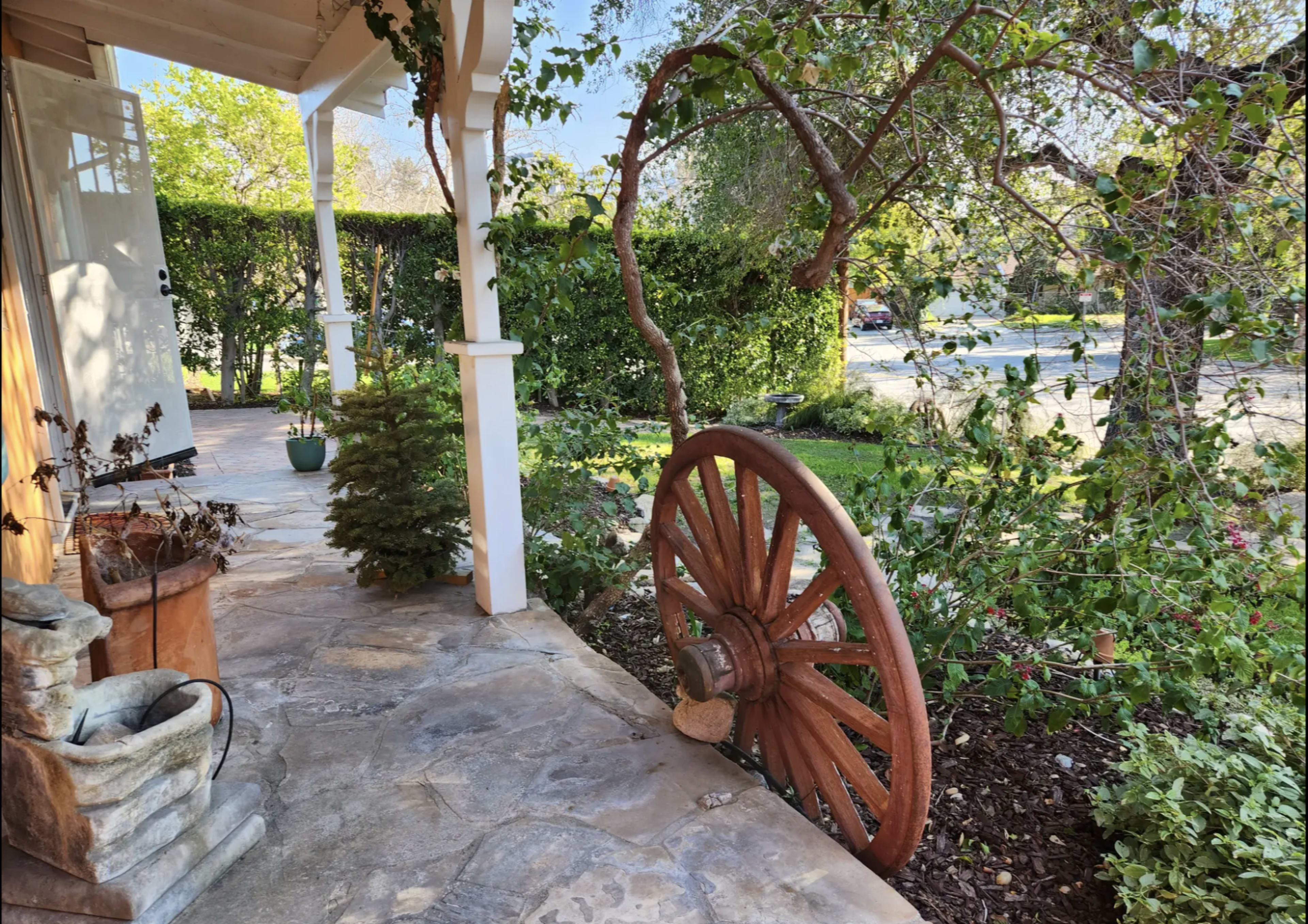The image shows a rustic porch area with a large wooden wagon wheel positioned beside a stone pathway and lush greenery.