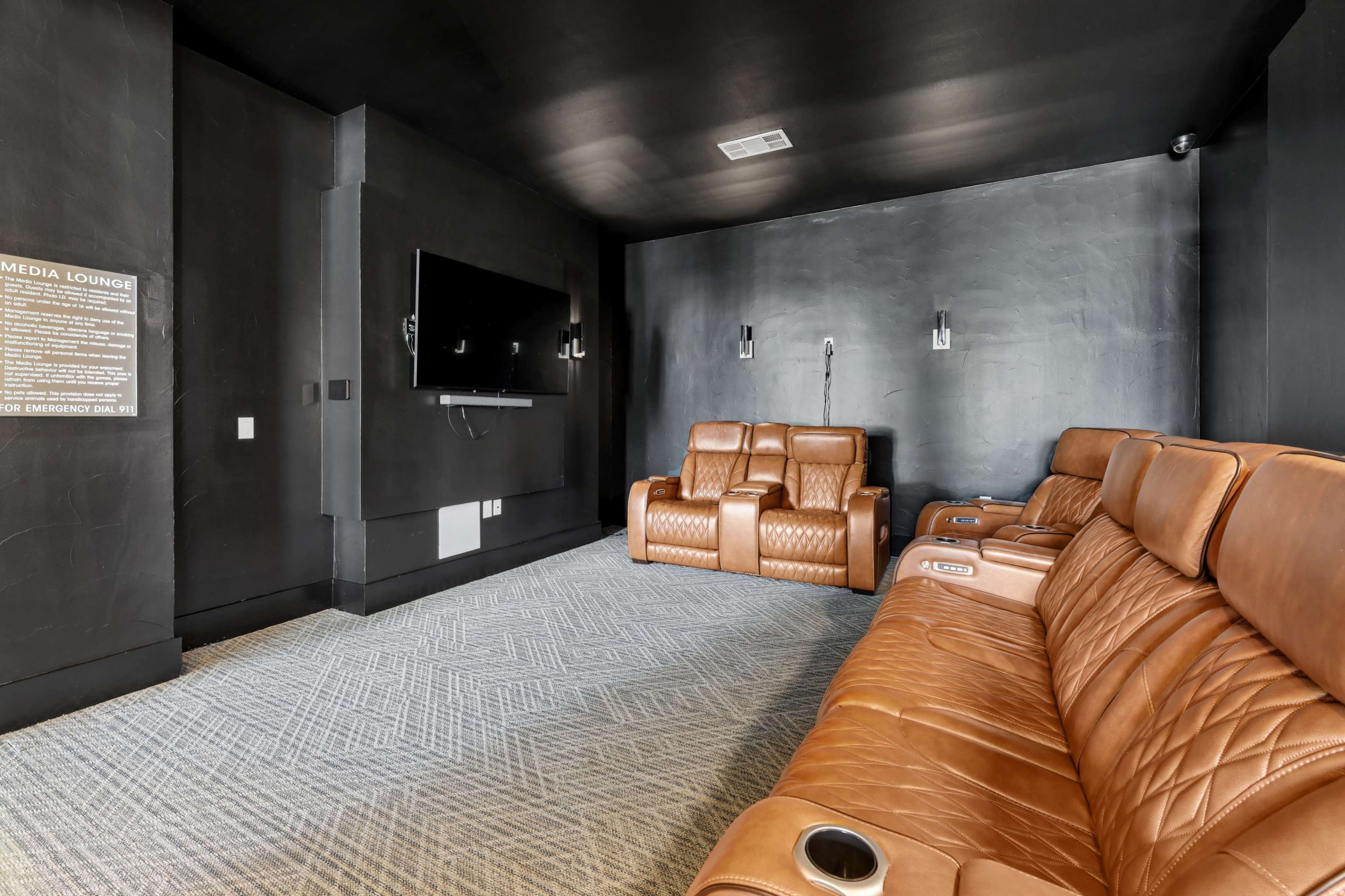 The image shows a modern media lounge with black walls, a large wall-mounted television, and brown leather recliner seating arranged on a textured carpet.