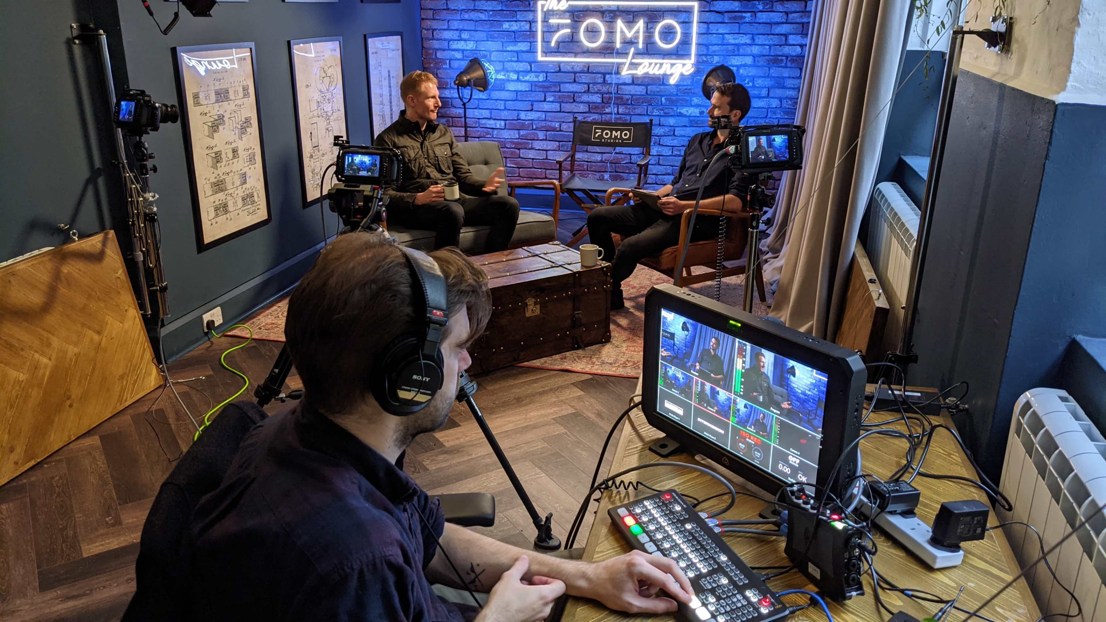 Two people are seated in a studio setting with a brick wall backdrop, discussing content while a third person operates a video switcher at a control desk.