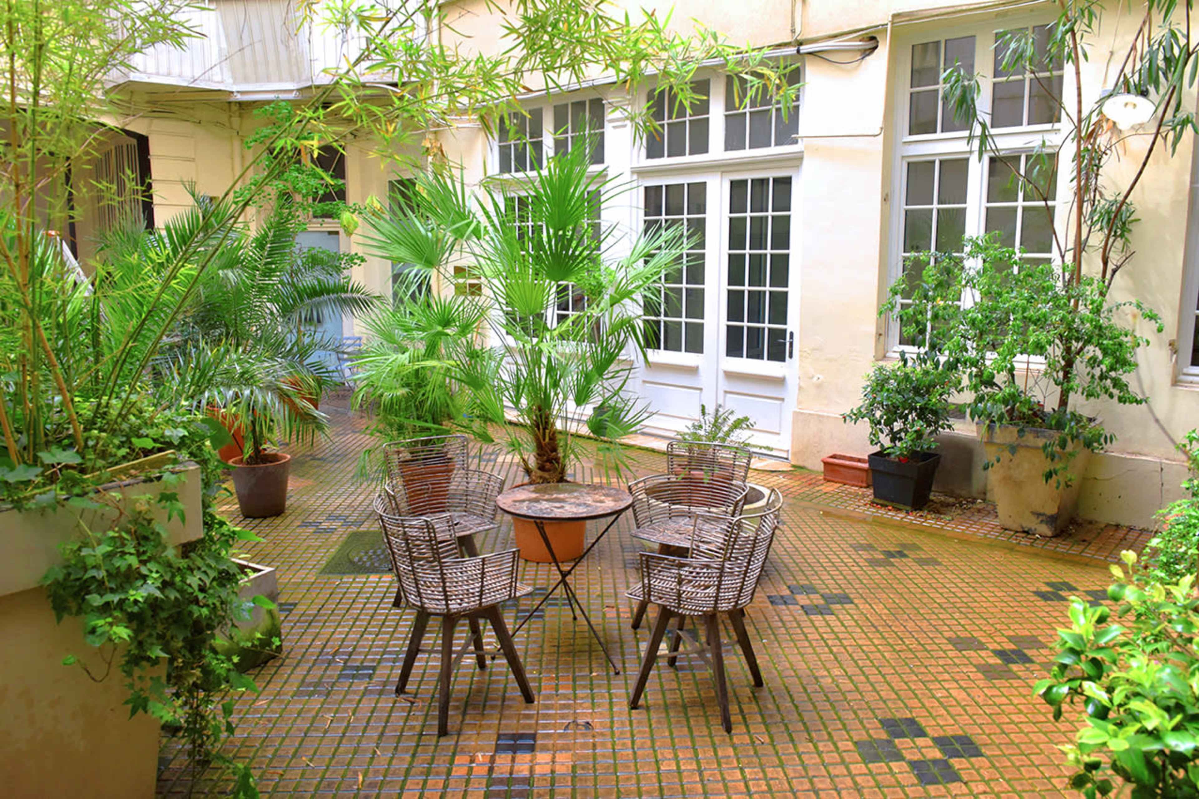 A small courtyard features a circular table surrounded by four woven chairs, set among various potted plants and greenery.