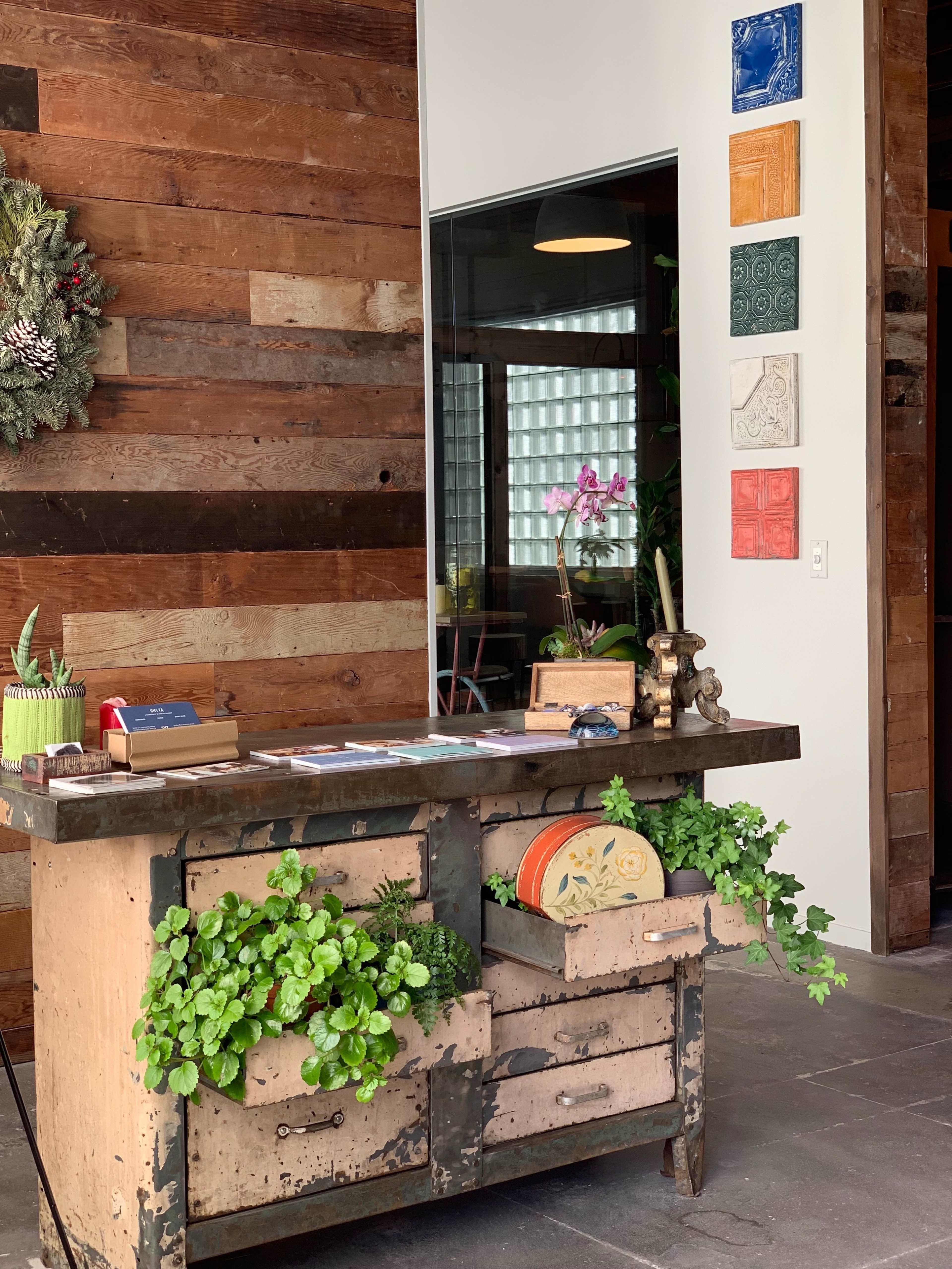 A wooden reception desk with plants, an orchid, and decorative items stands against a wall with wooden paneling and framed art pieces.