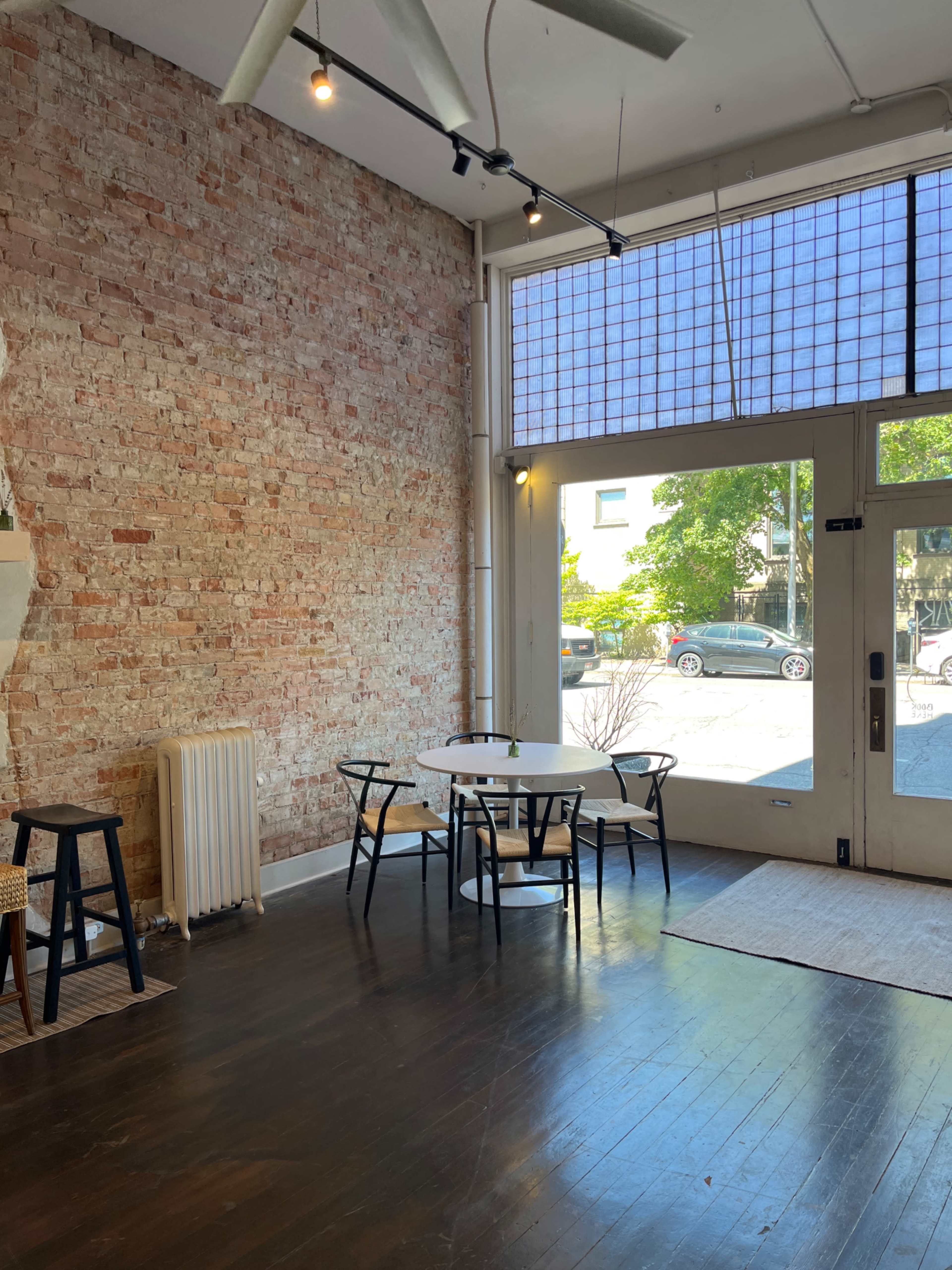 A spacious interior with a brick wall, a round table surrounded by four chairs, and a large window that opens to a street view.
