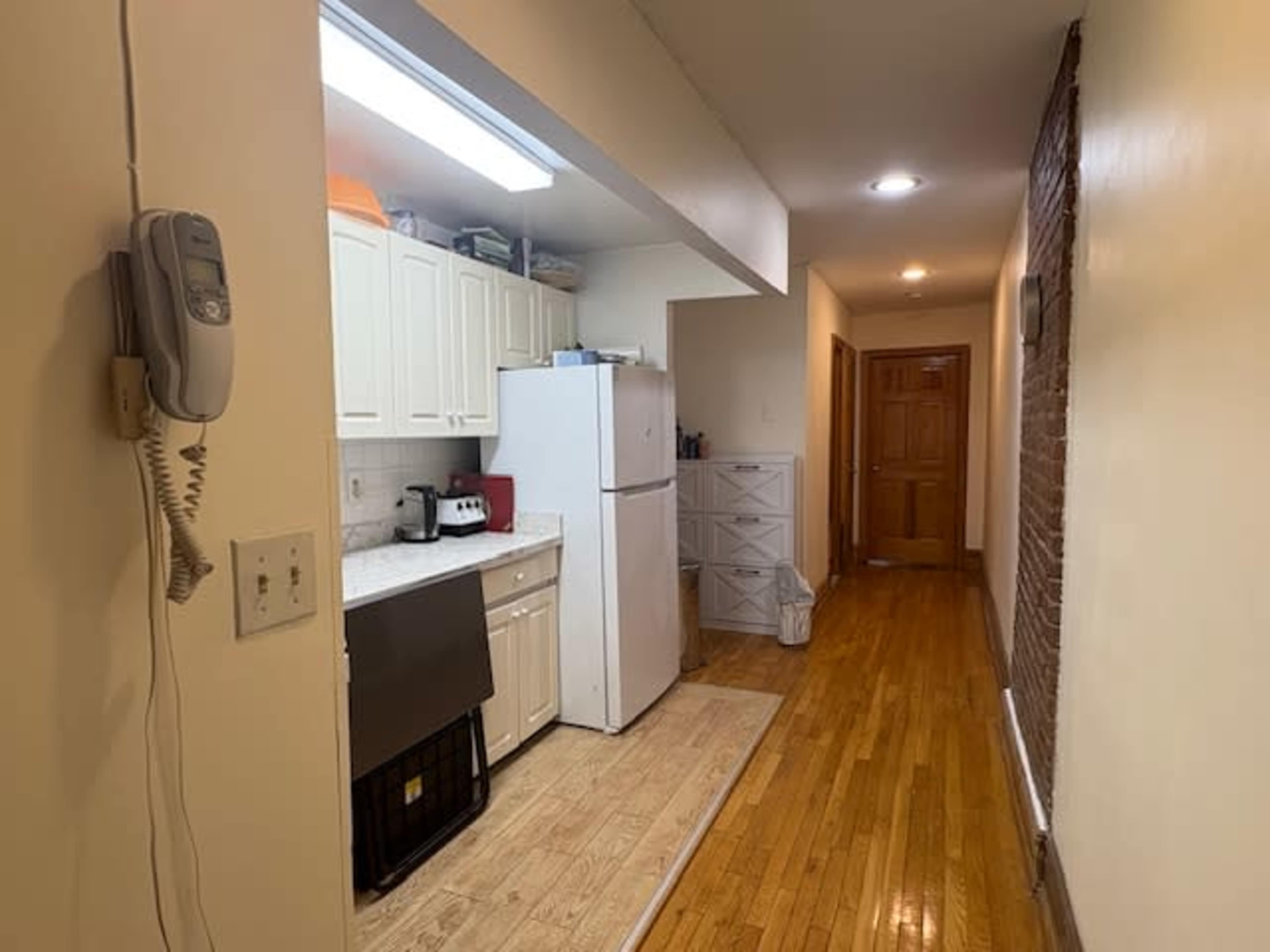 The image shows a narrow hallway leading to a kitchen area, featuring white cabinetry, a refrigerator, and wood flooring.