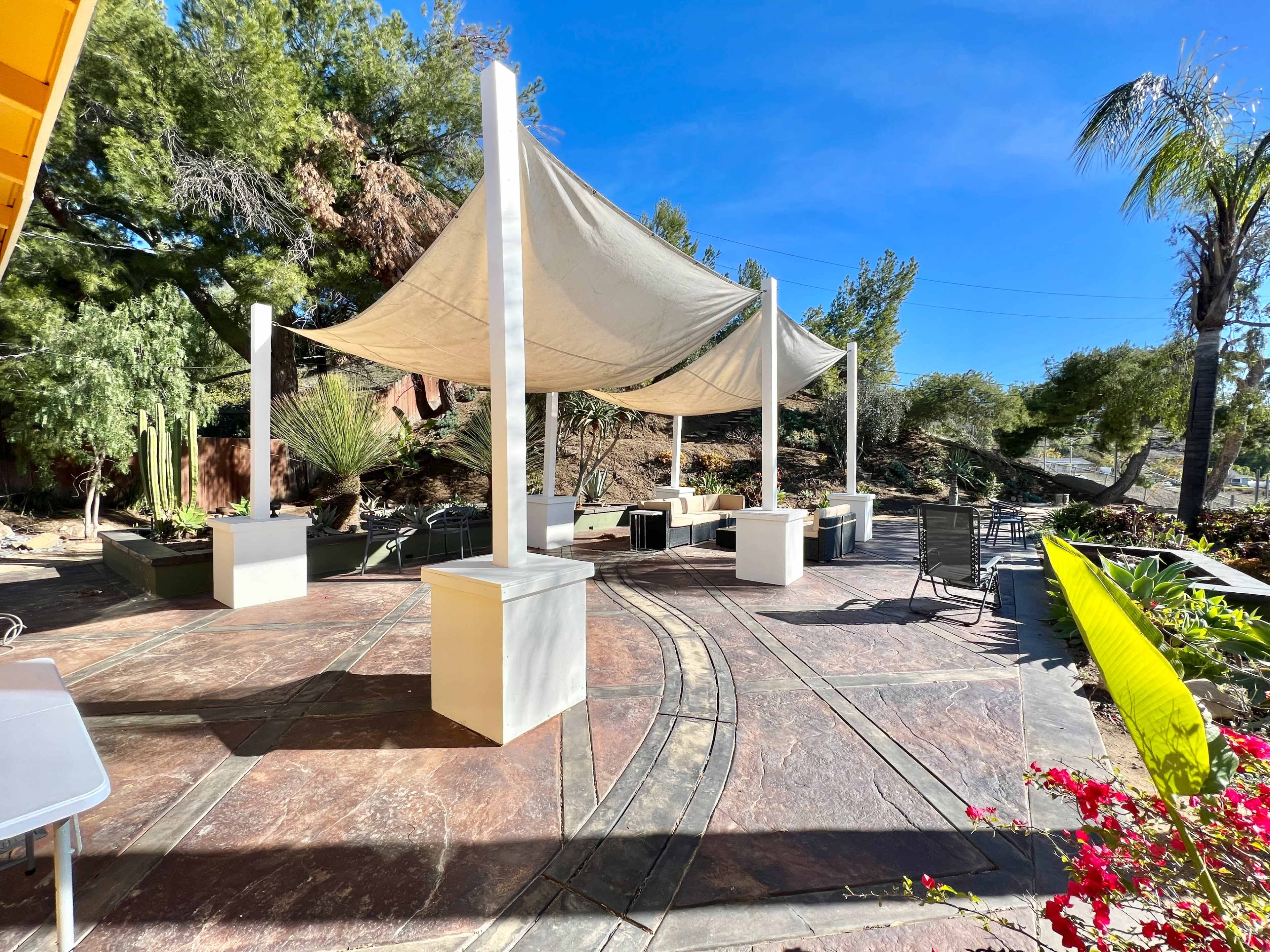 A patio area with shaded seating under a canopy, surrounded by landscaping and palm trees.