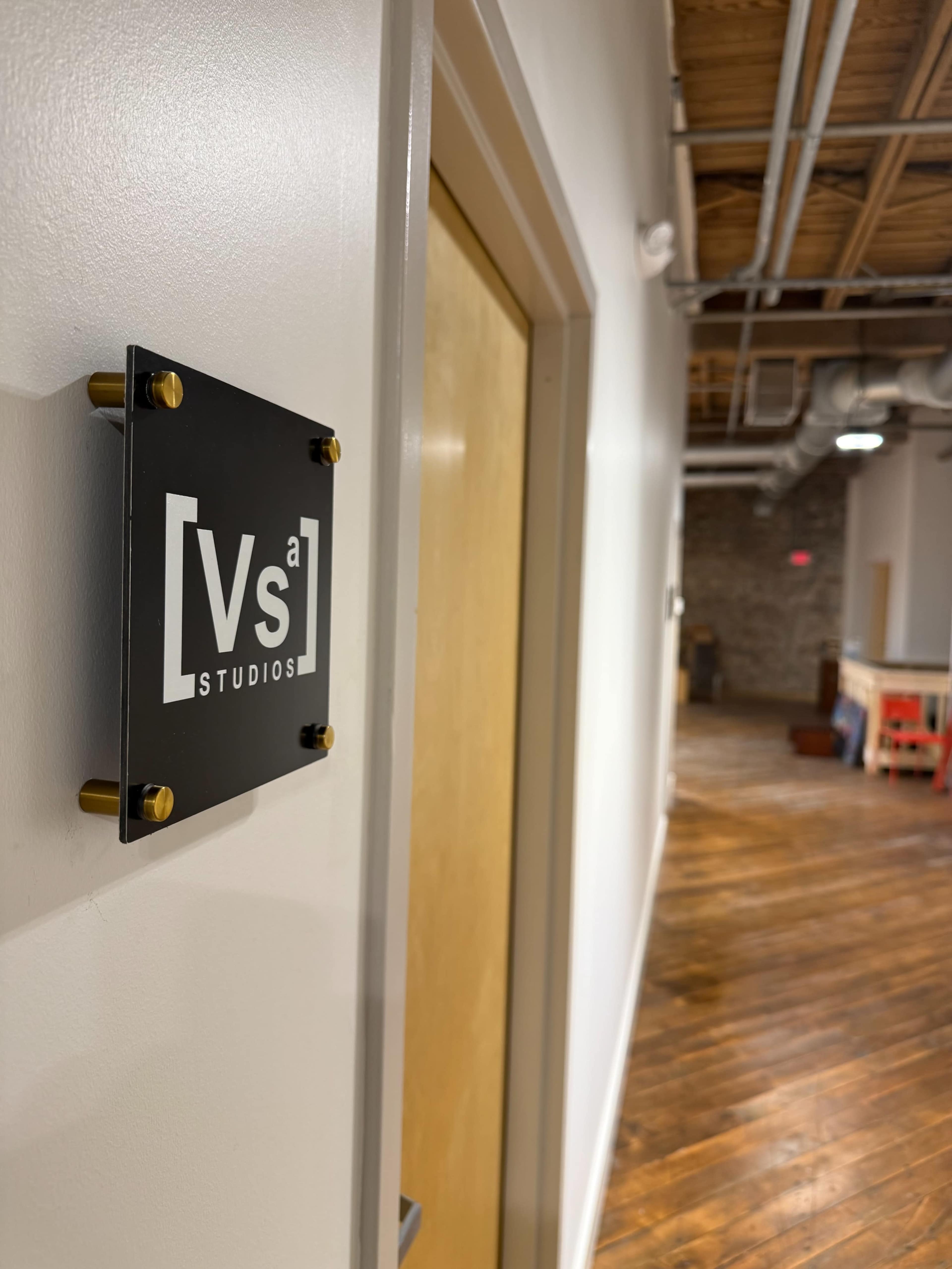 A sign indicating "Vs[a] STUDIOS" is attached to a door in a well-lit hallway with wooden flooring and exposed ceiling.
