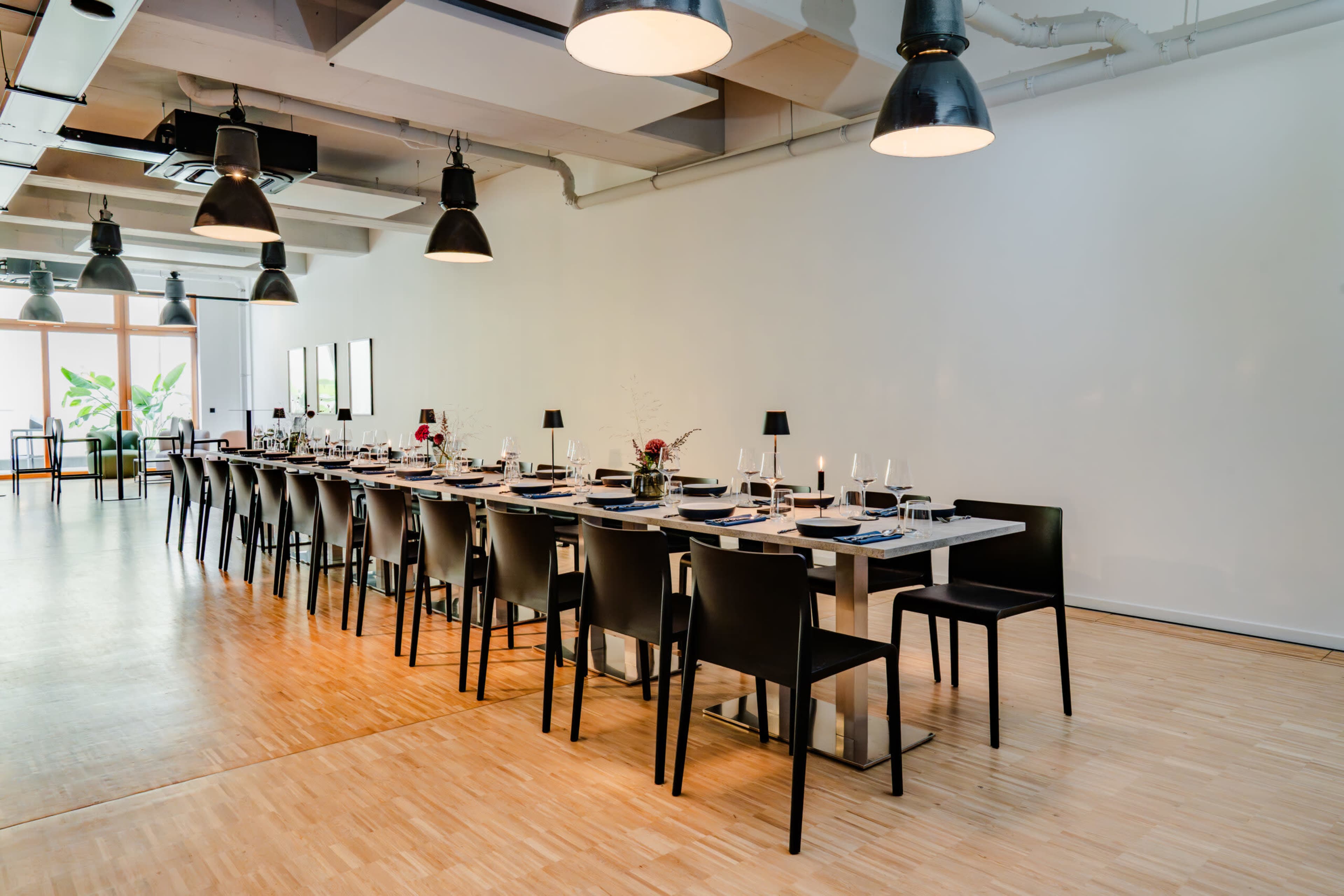 A long dining table is set with plates, glasses, and cutlery, surrounded by black chairs in a well-lit, spacious room.