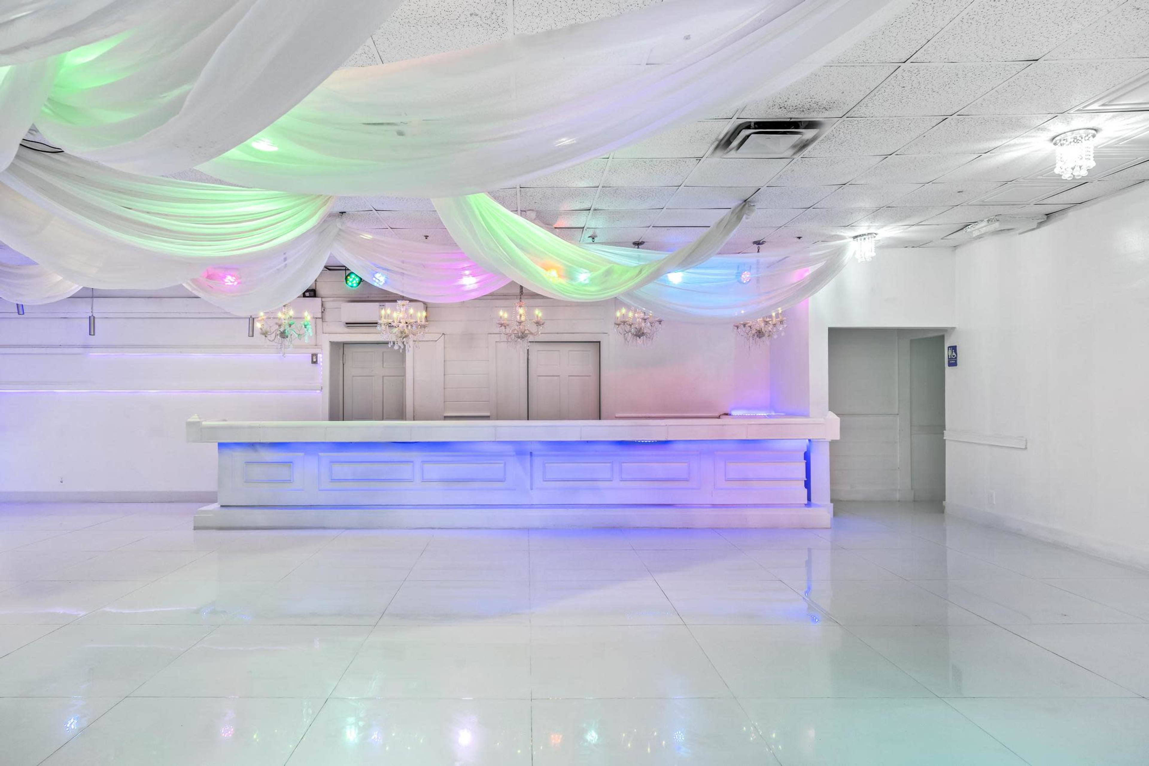 The image shows a spacious event venue with a white backdrop, featuring a bar area illuminated by colorful lights and draped fabric overhead.