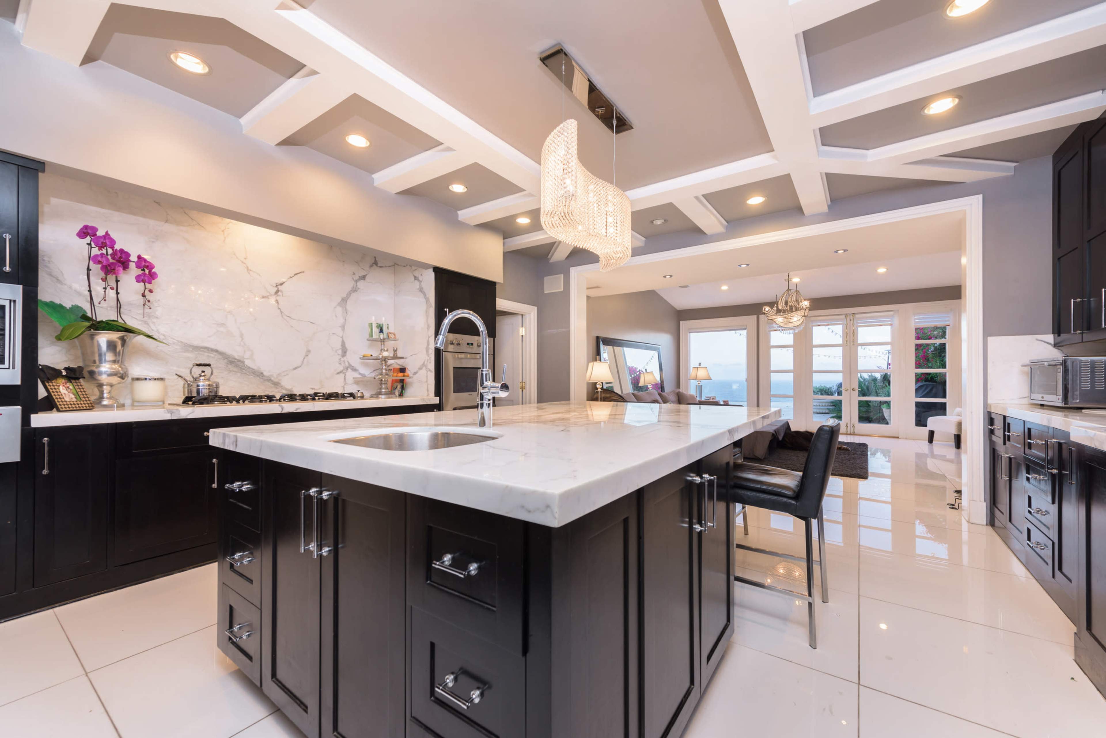 The image shows a modern kitchen featuring a large marble island, dark cabinetry, and an elegant chandelier above.