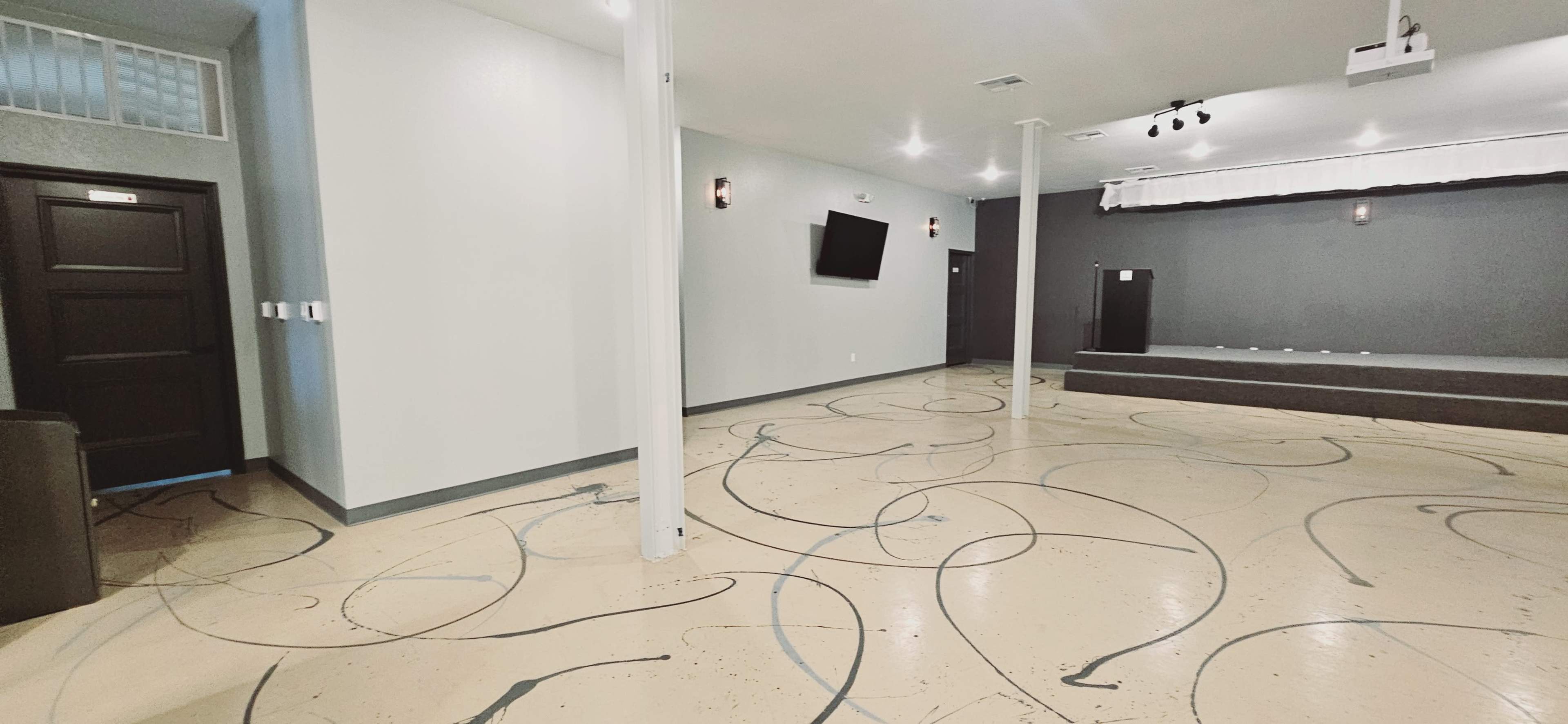 An empty interior space with a smooth, patterned floor, dark walls, a small stage, and a wall-mounted television.