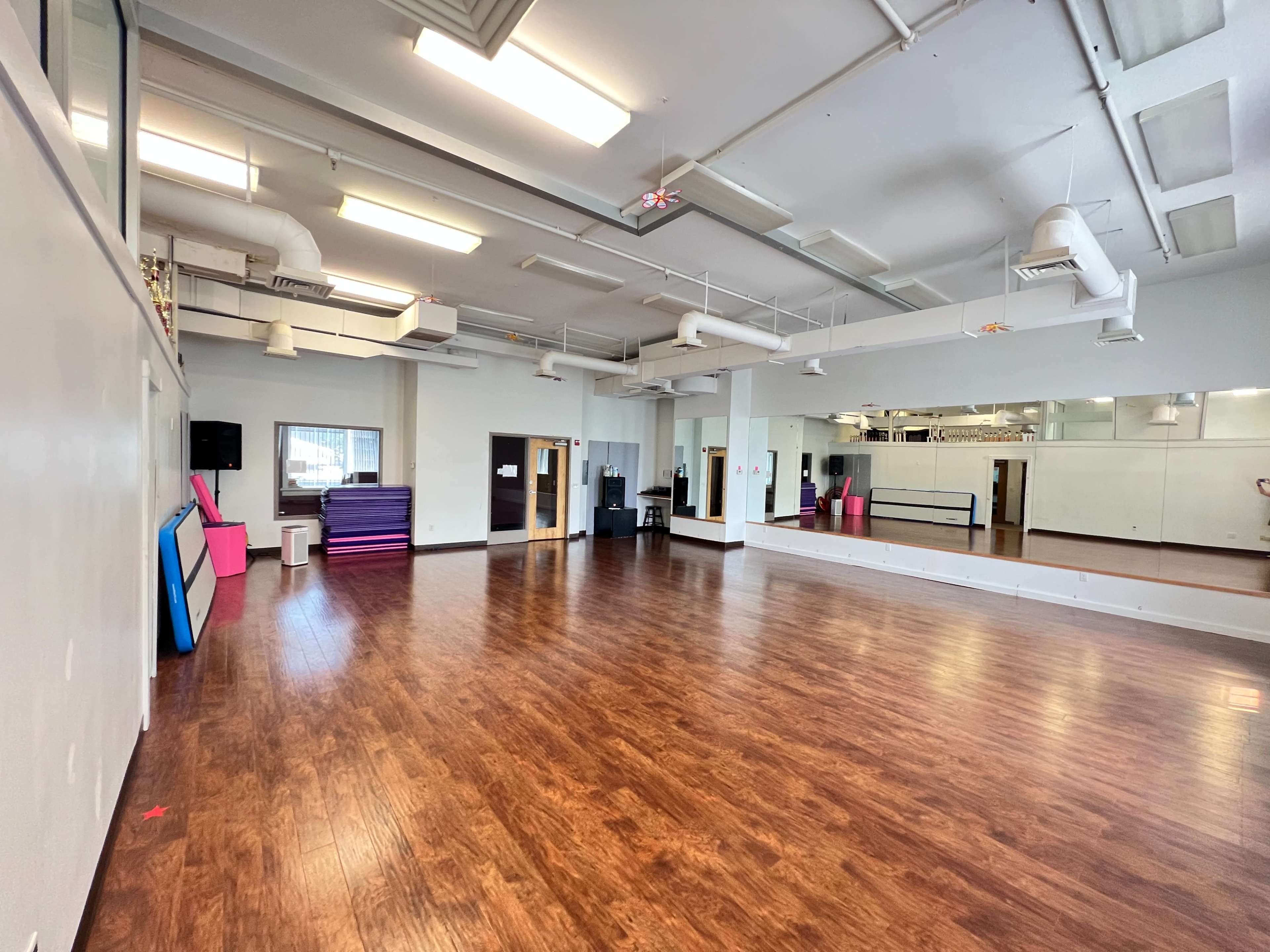 A spacious dance studio with hardwood flooring, mirrors on one wall, and colorful mats stacked in the corner.